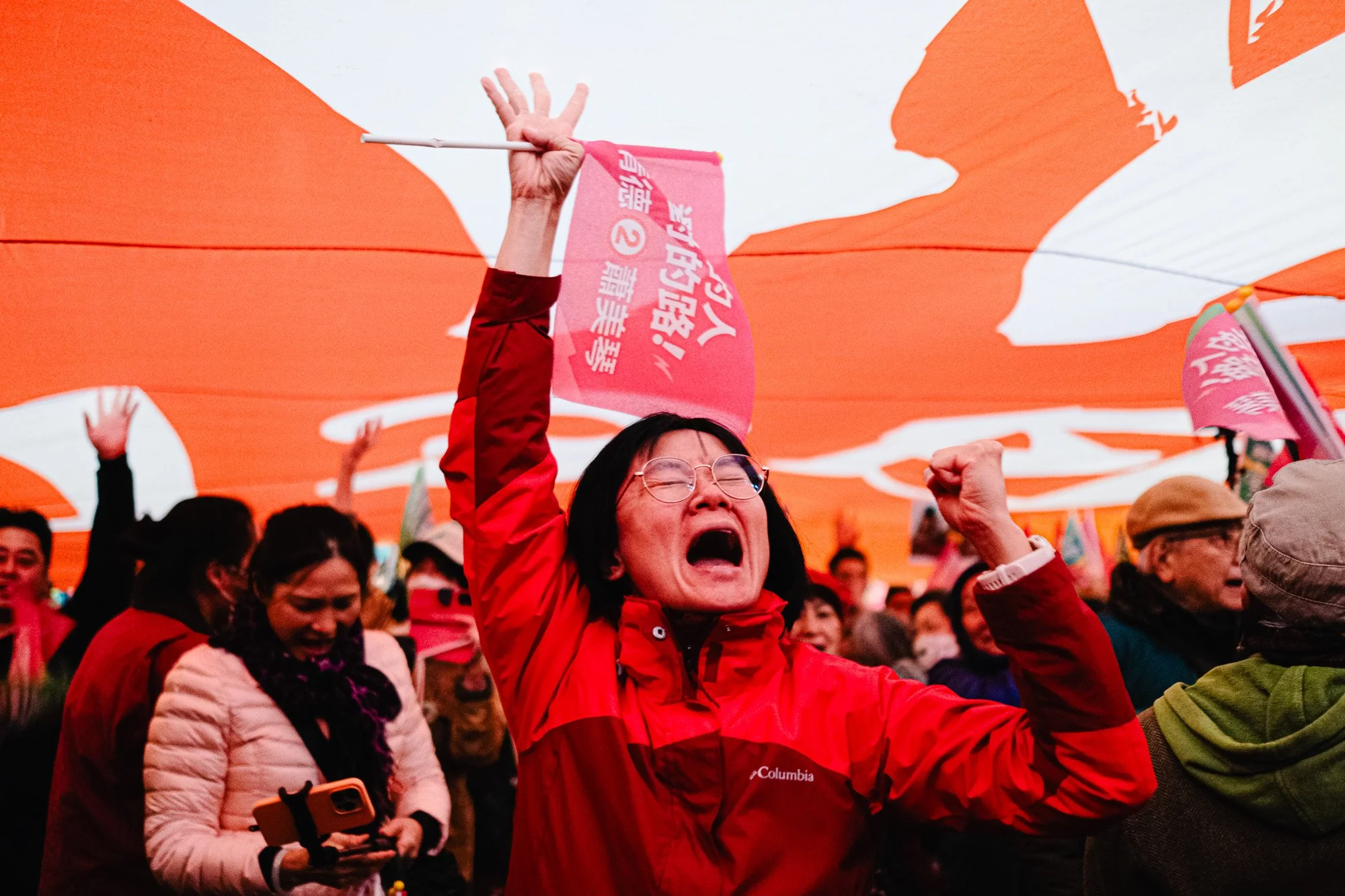 A group of people celebrating outdoors, with a woman in a red Columbia jacket holding a pink flag and cheering. The background features orange umbrellas and a crowd of diverse individuals.