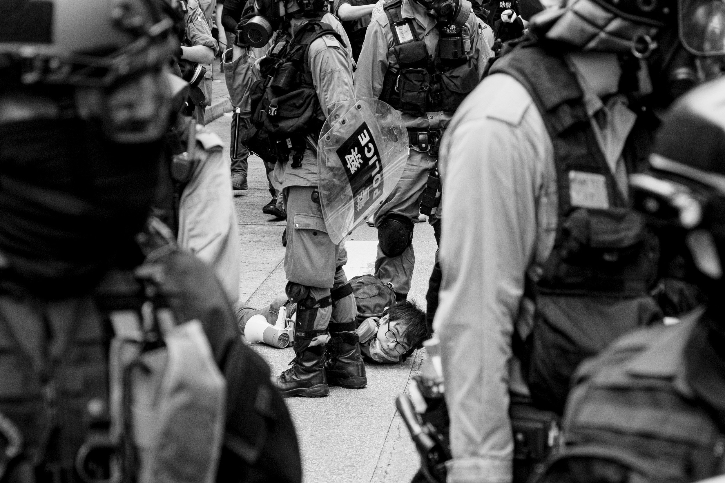 Black and white photo of police officers in uniform with riot gear, forming a line on a street. A person lying on the ground is visible between officer legs, wearing glasses and a mask, with a water bottle nearby.