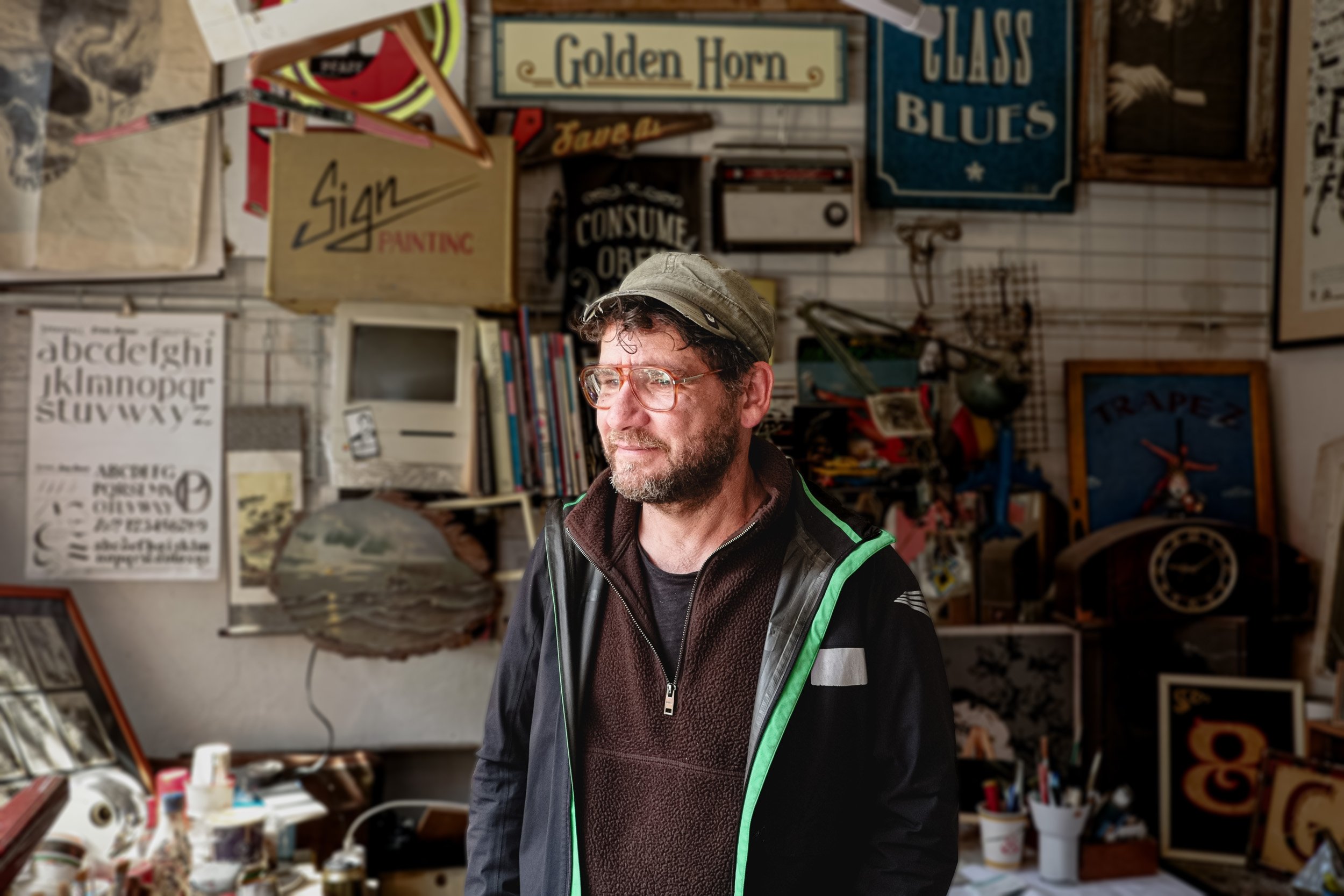 A man with glasses and a beige cap standing in a craft or antique shop with various signs and items on the walls behind him.