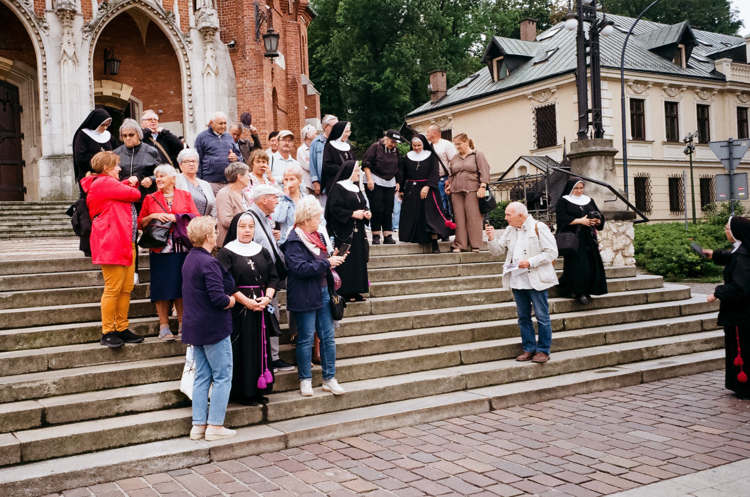 Group of people, including nuns in black habits and laypeople, gathered on the stairs in front of a historic building with brick and stone architecture and trees in the background.