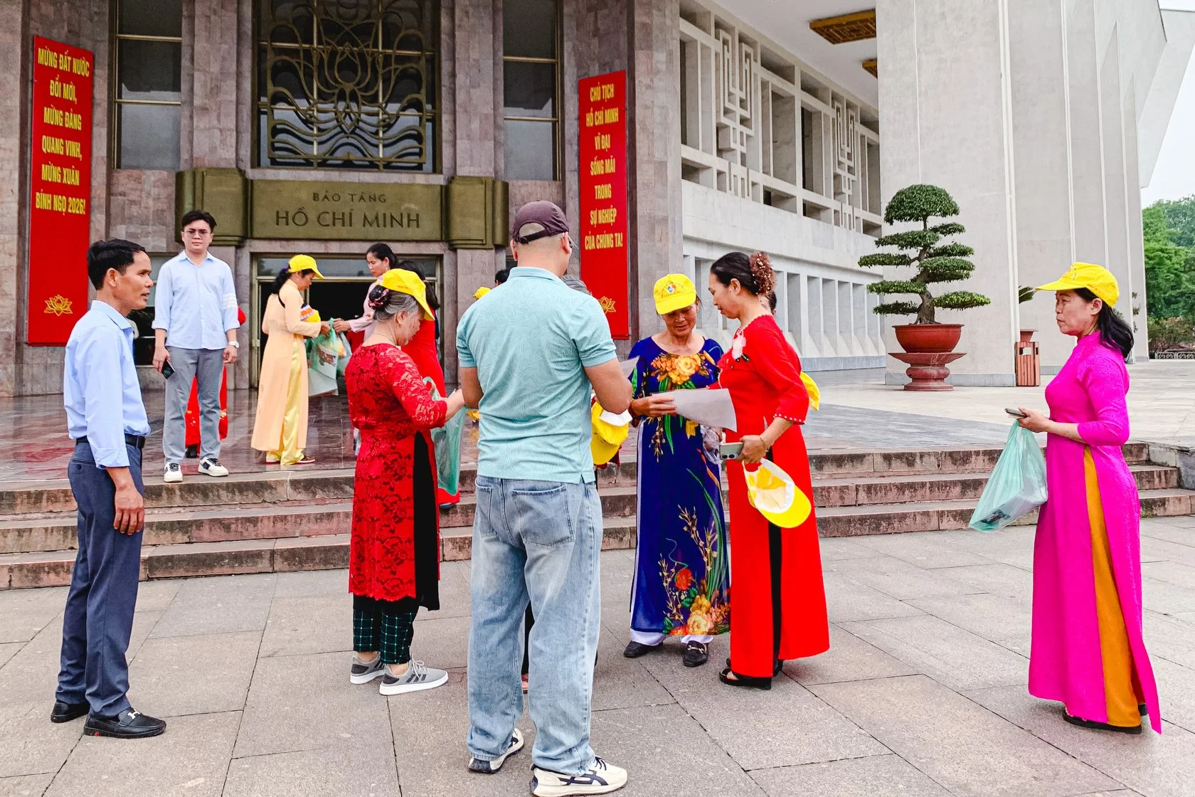 Group of people gathered outside the Ho Chi Minh Museum, some in traditional Vietnamese attire, engaging in conversation and reading papers, with the museum building and potted plant in the background.