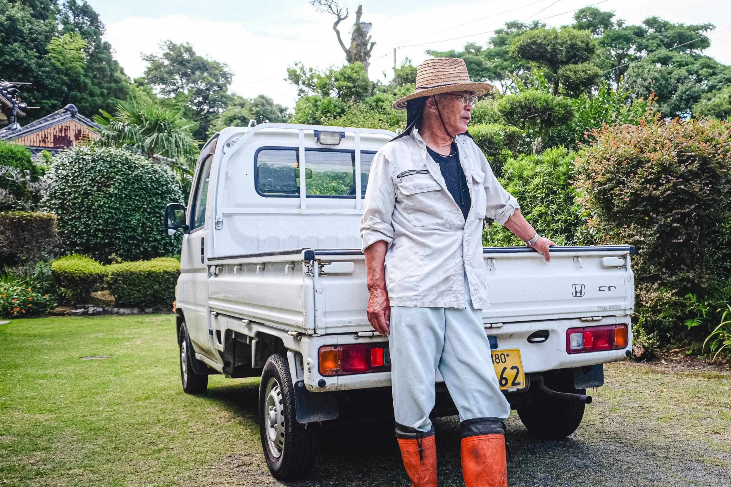 An elderly man wearing a large straw hat, glasses, and work clothes stands next to a small white Honda pickup truck in a garden with lush green trees and bushes.