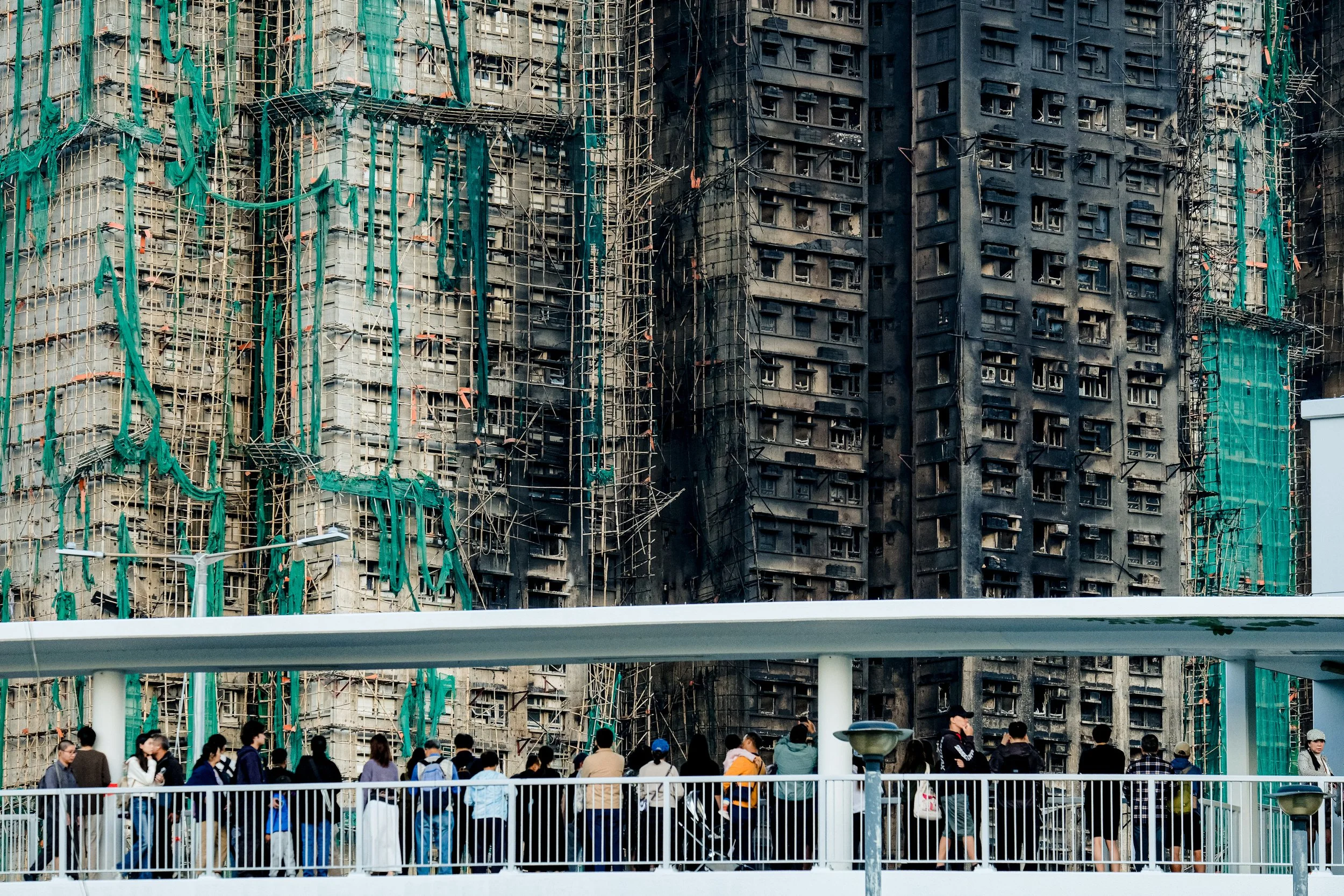People are gathered in front of a tall building under construction, which is covered in scaffolding and green construction netting.