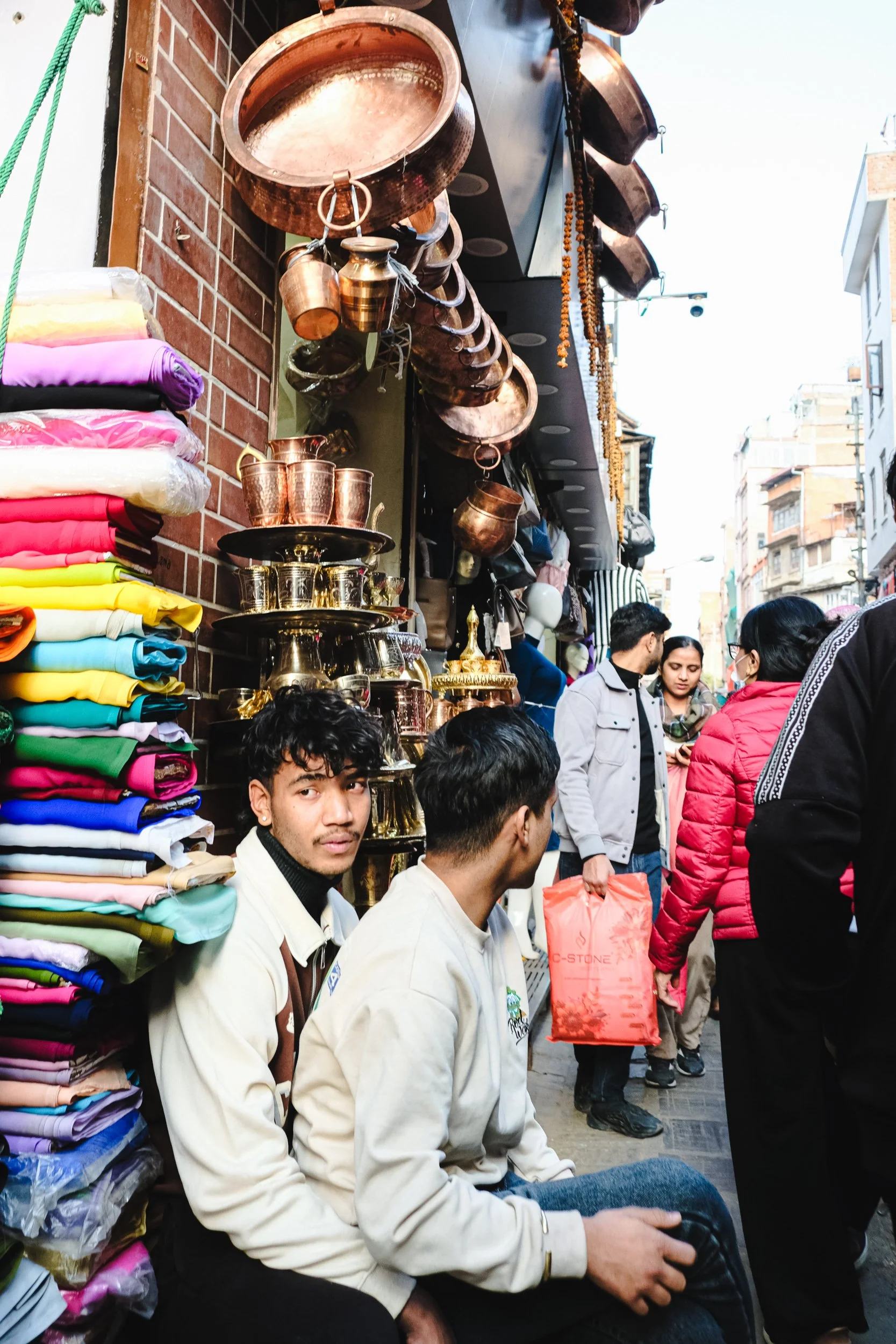 People shopping at an outdoor market with colorful clothes on shelves to the left, hanging copper cookware on the wall, and many shoppers walking along a narrow street lined with buildings.