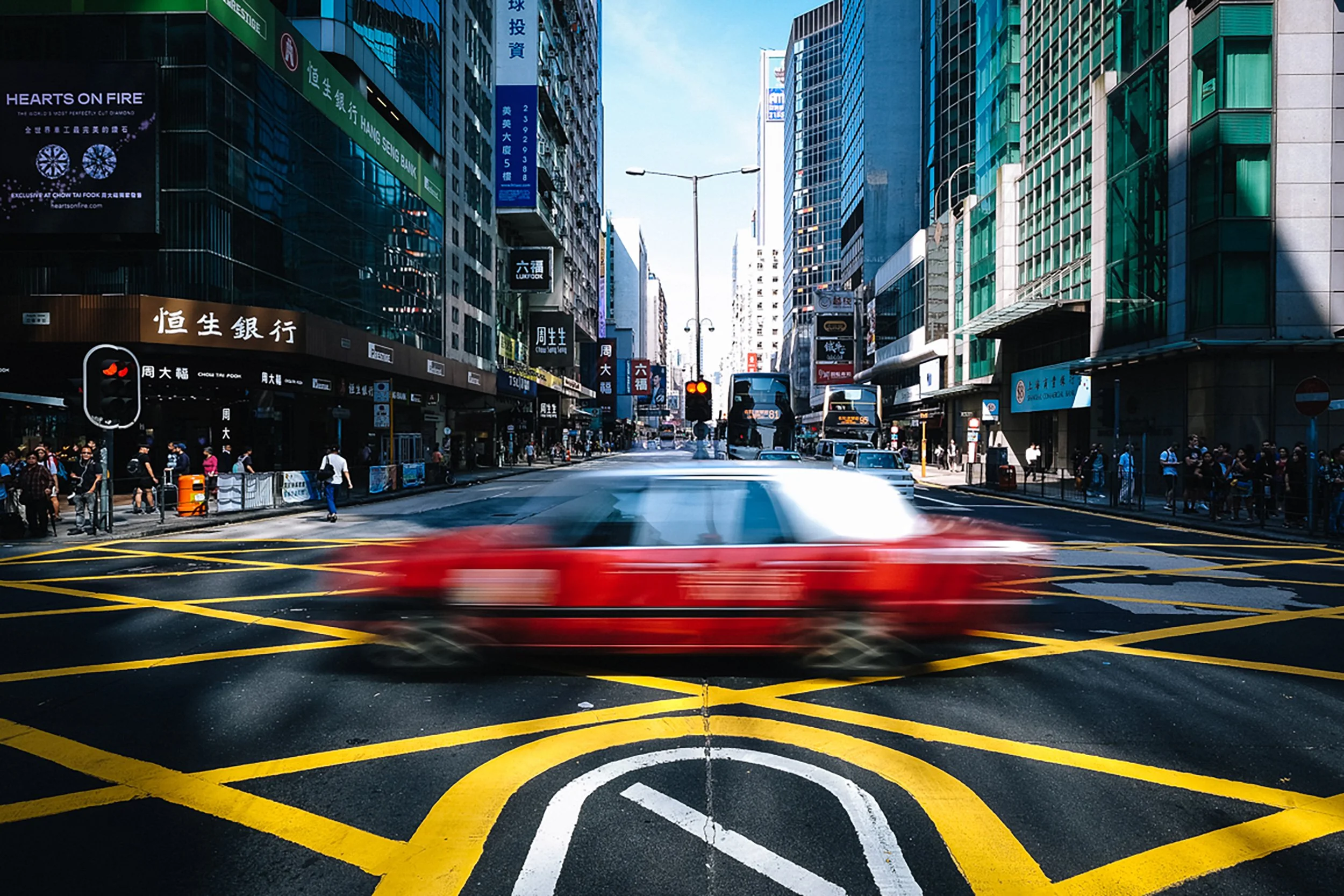 A busy city street with tall glass buildings, pedestrians crossing, and vehicles, including a red car in motion, captured during daytime with a clear sky.