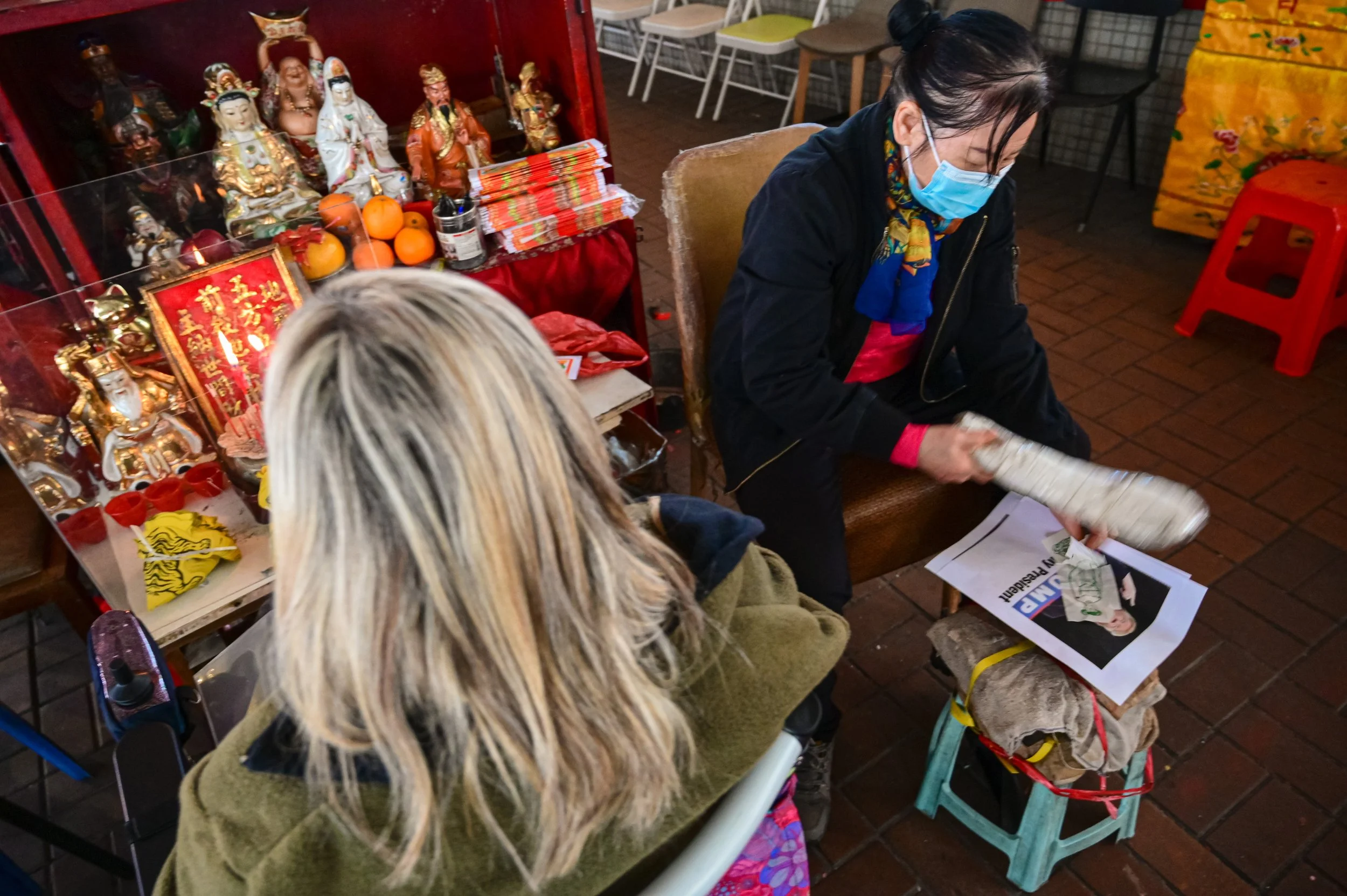 A woman wearing a blue face mask and dark jacket is seated at a table, looking down at documents. Behind her is a display of various figurines and offerings, including oranges, religious statues, and candles, indicating a cultural or religious settin
