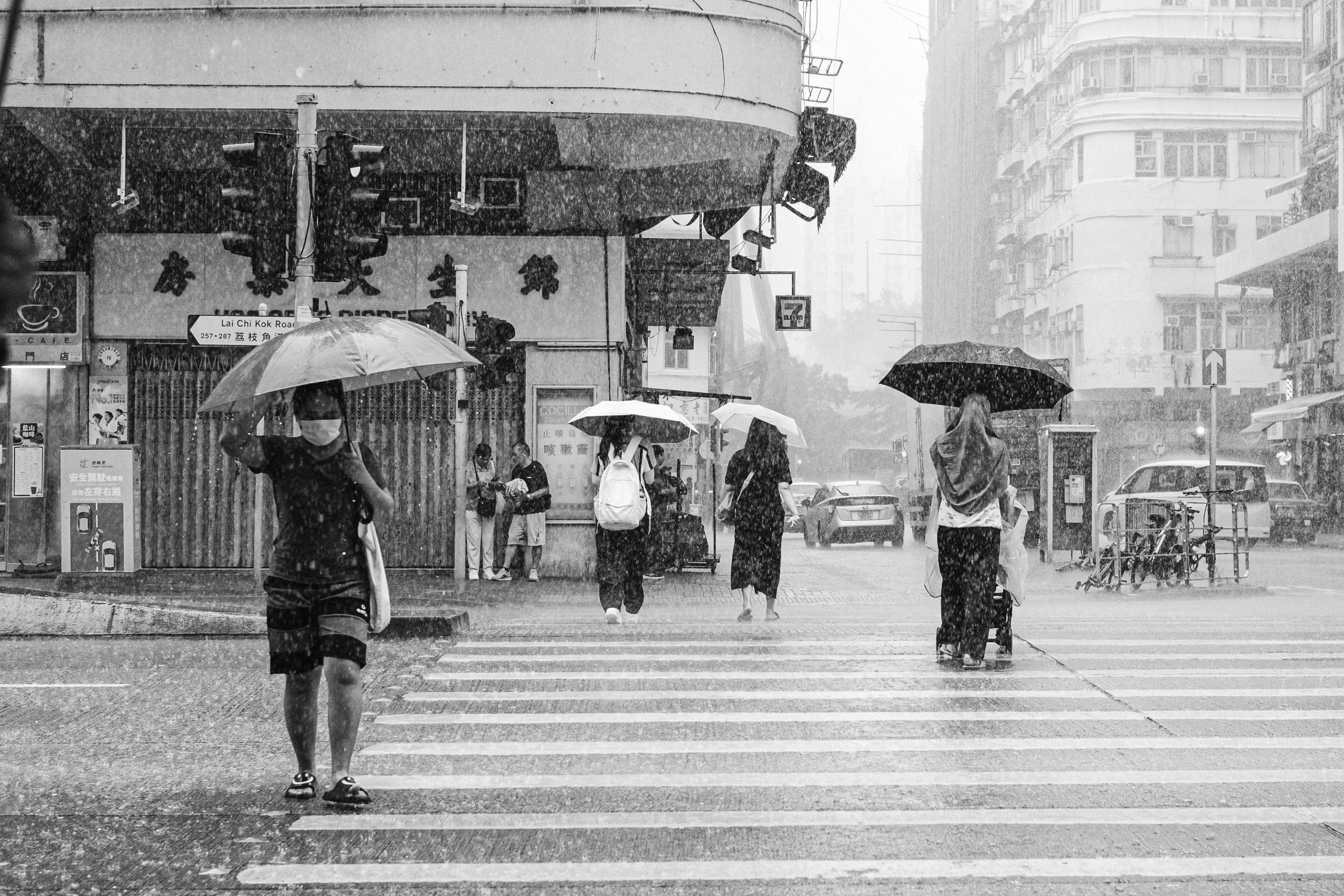 People walking across a rainy city intersection with umbrellas, several people waiting on the sidewalk, tall buildings, and a storefront with Chinese characters.