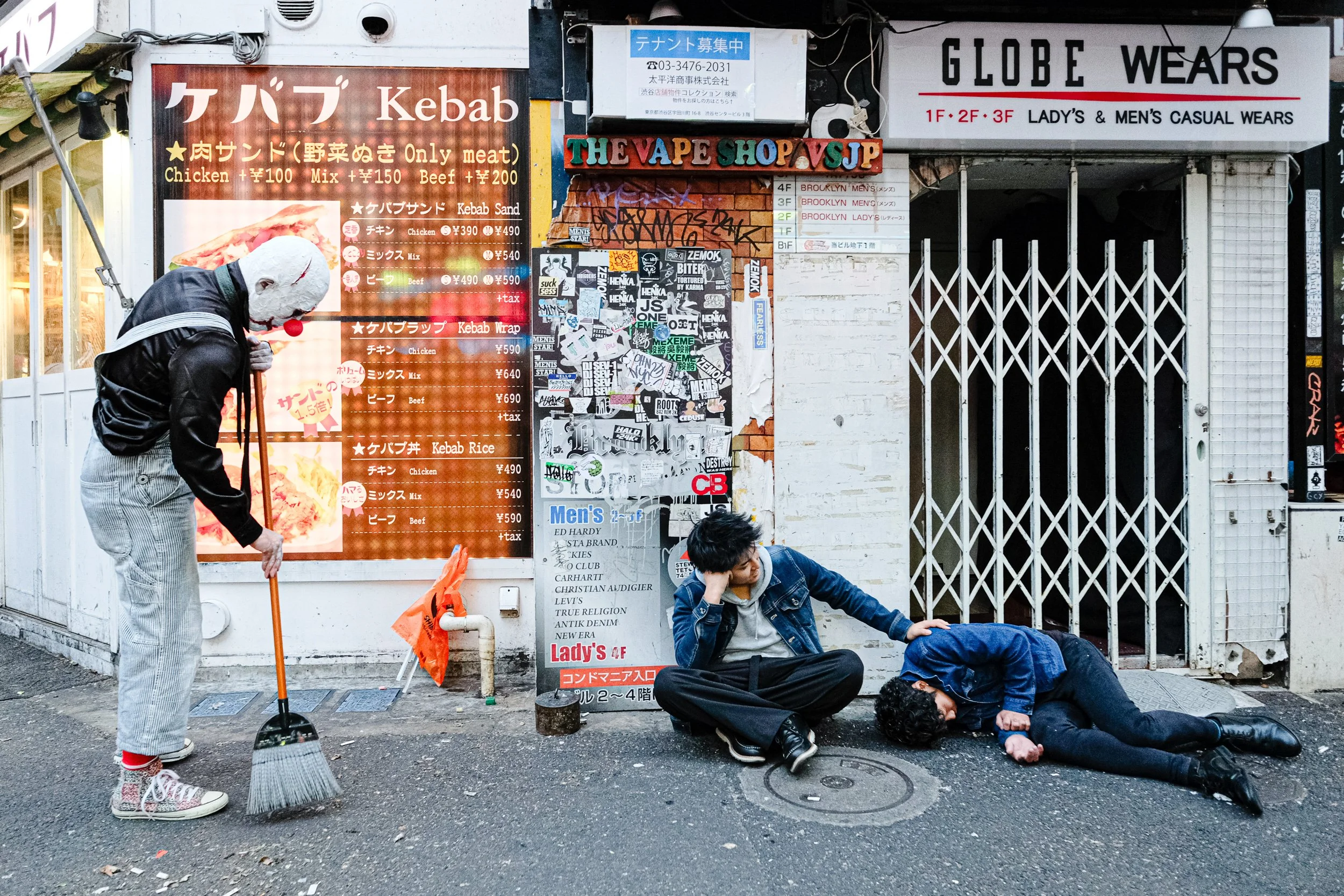 Street scene with two young men, one lying on the ground, the other sitting and leaning on his elbow, in front of a closed shop with graffiti and signage. A person with a mask and casual clothing is sweeping the sidewalk nearby.