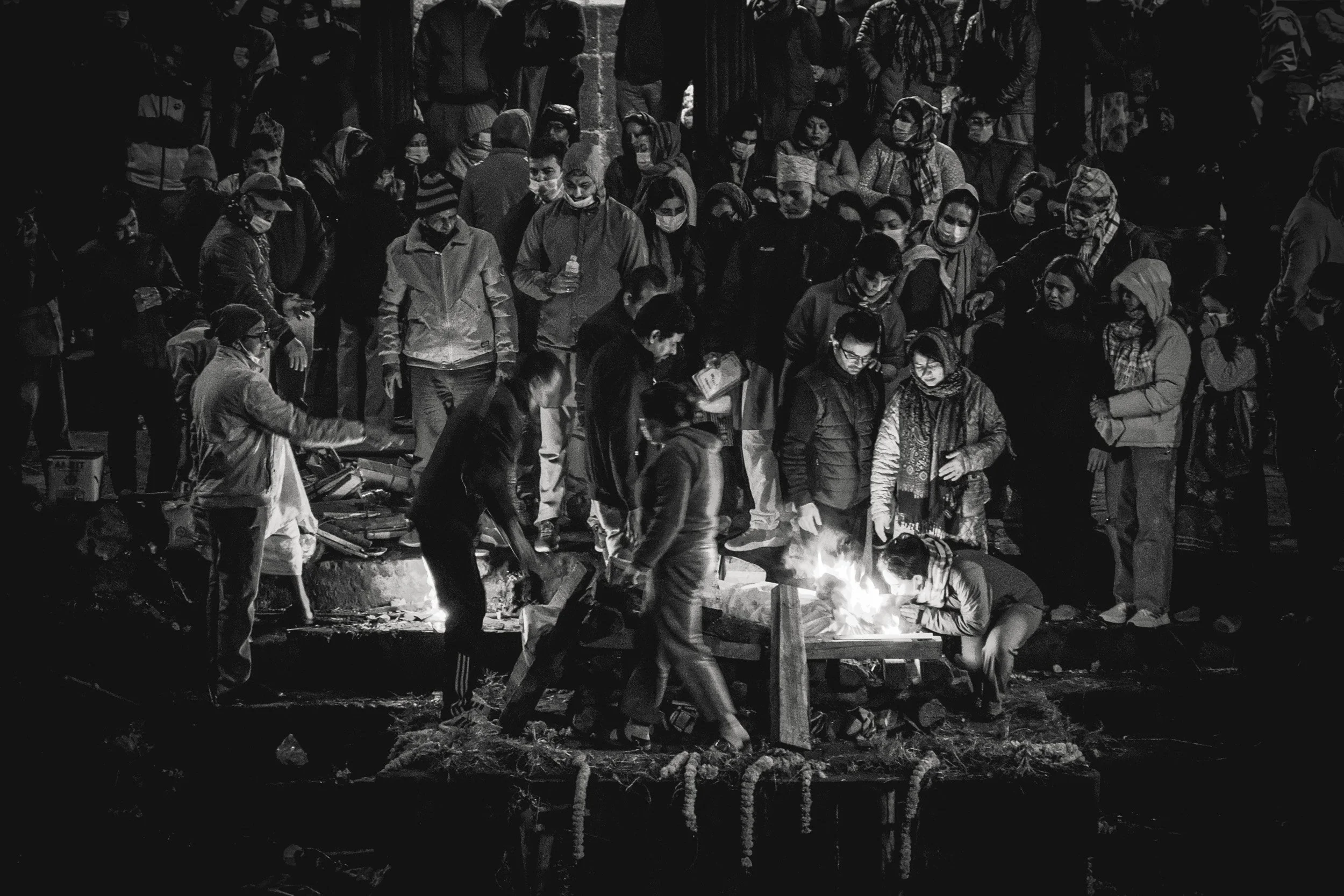 A large group of people gathered outdoors at night, many wearing masks. Some are standing, while others are sitting or working around a fire on the ground. The scene appears to be lively and crowded, with a dark background and some visible faces and 