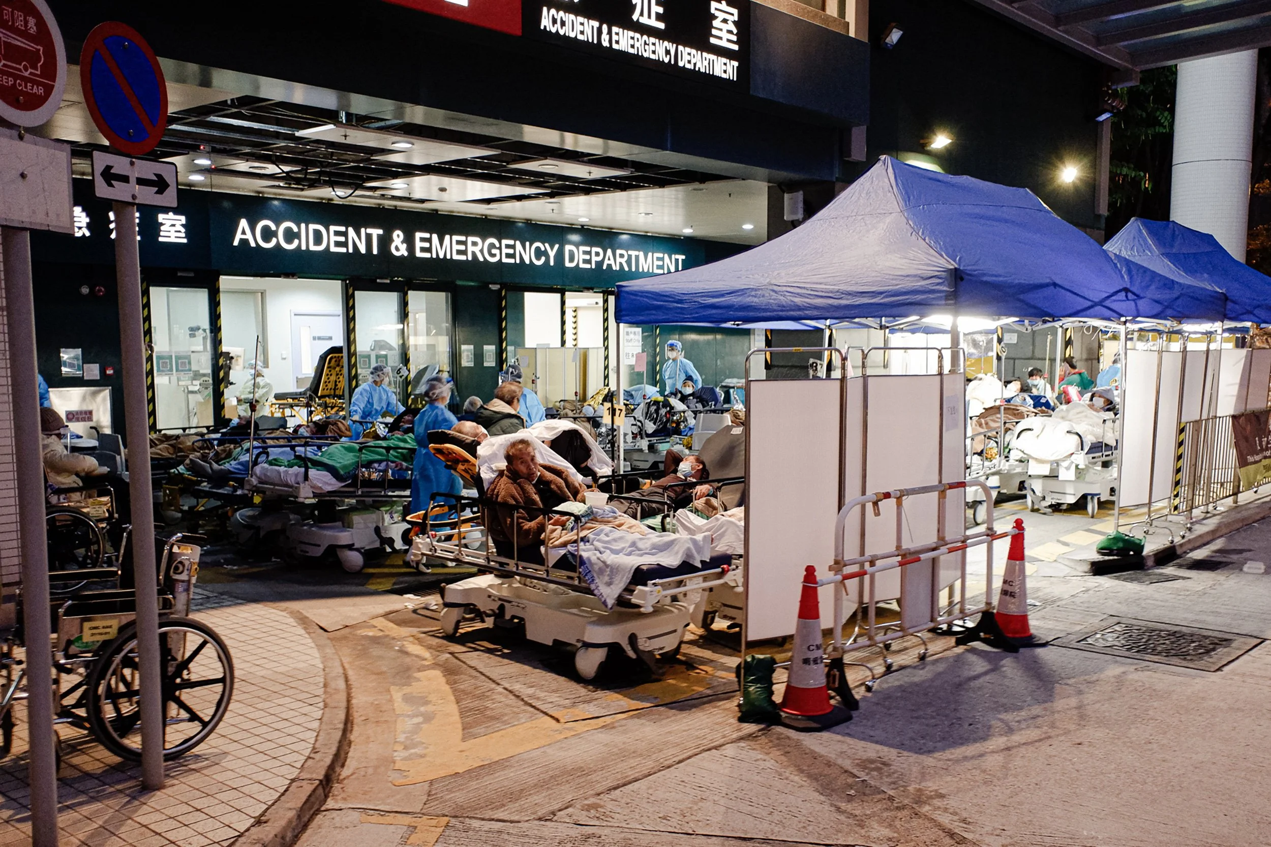 Nighttime scene of an outdoor hospital emergency area with numerous patients on stretchers under blue tents, medical staff, and traffic cones.