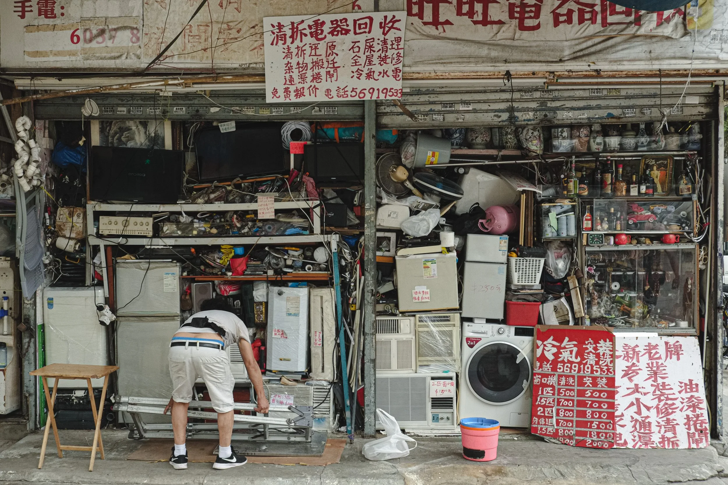 A person in white shorts and black sneakers is fixing a metal gate in front of a cluttered storefront. The shop has various electronics, appliances, and miscellaneous items stacked and arranged inside. Red and white signs with Chinese characters and 