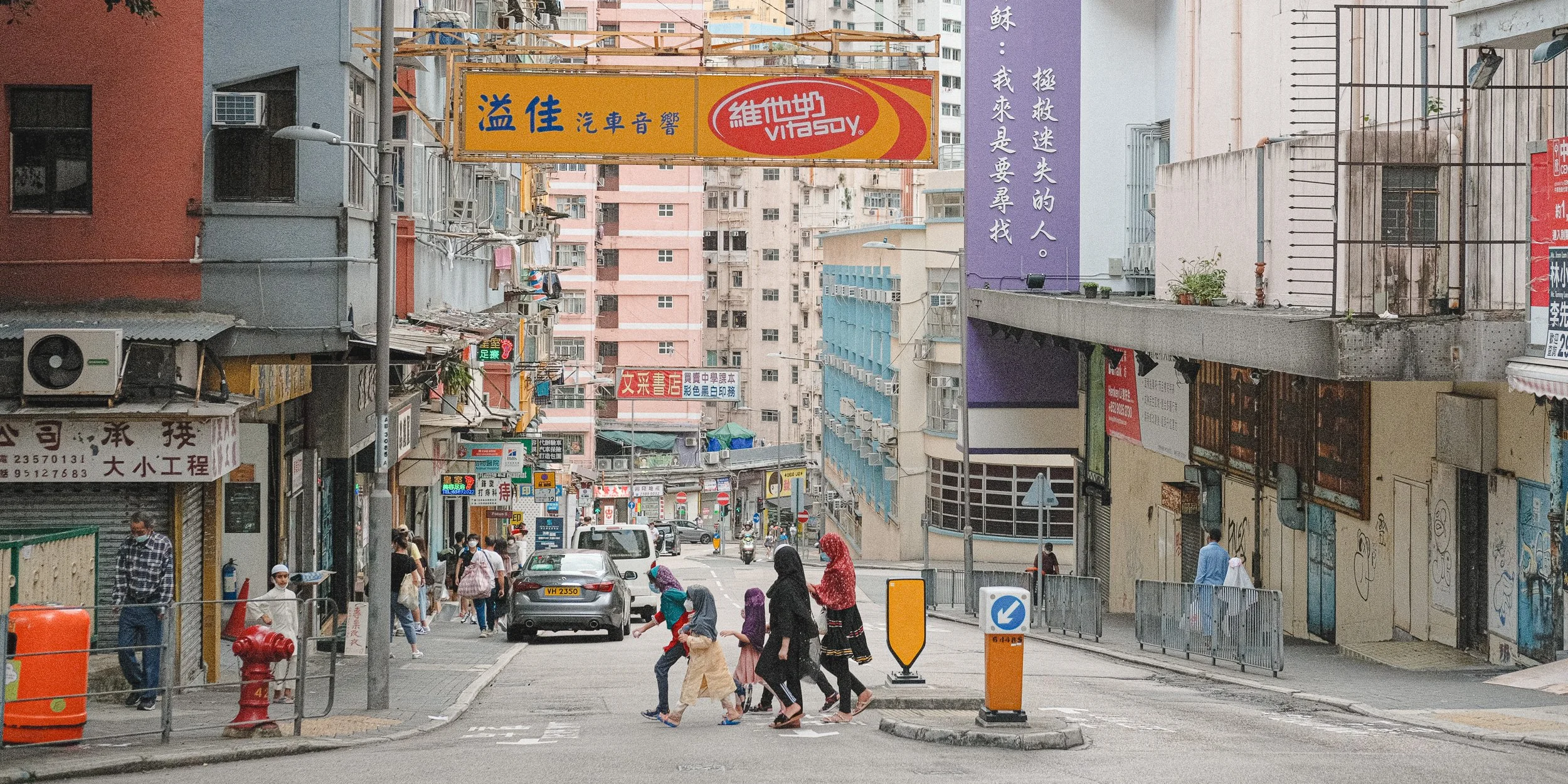 Street scene with pedestrians crossing and vehicles on the road, surrounded by multi-story buildings with various signs and advertisements in an urban area.