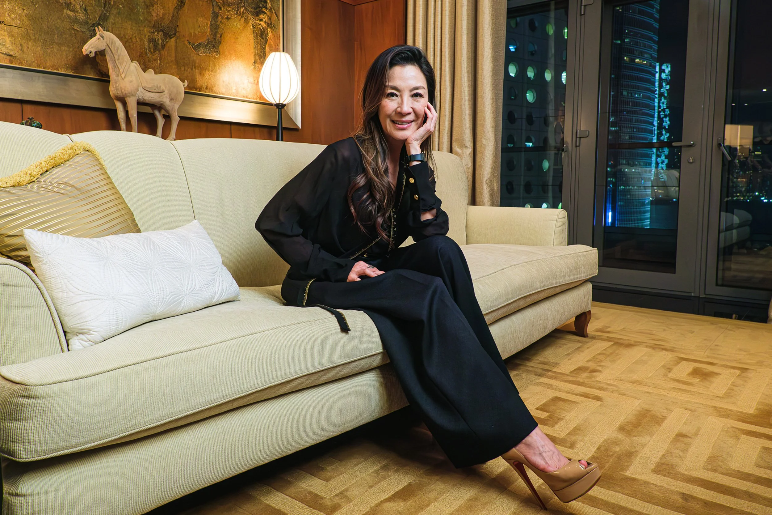 A woman sitting on a beige sofa in a modern apartment at night, with view of city lights outside the window. The room has wooden floor, table lamp beside her, and decorative horse sculpture on the wall.