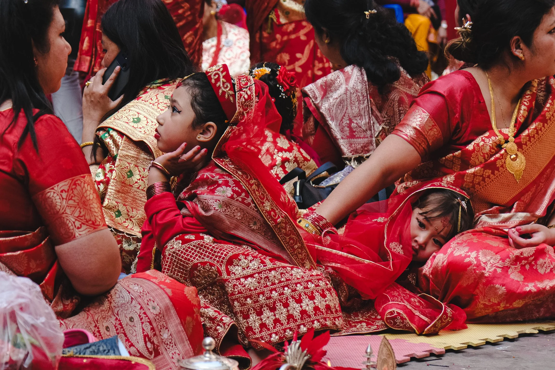 Women and children dressed in red and gold traditional attire, seated closely together at a cultural event or ceremony.