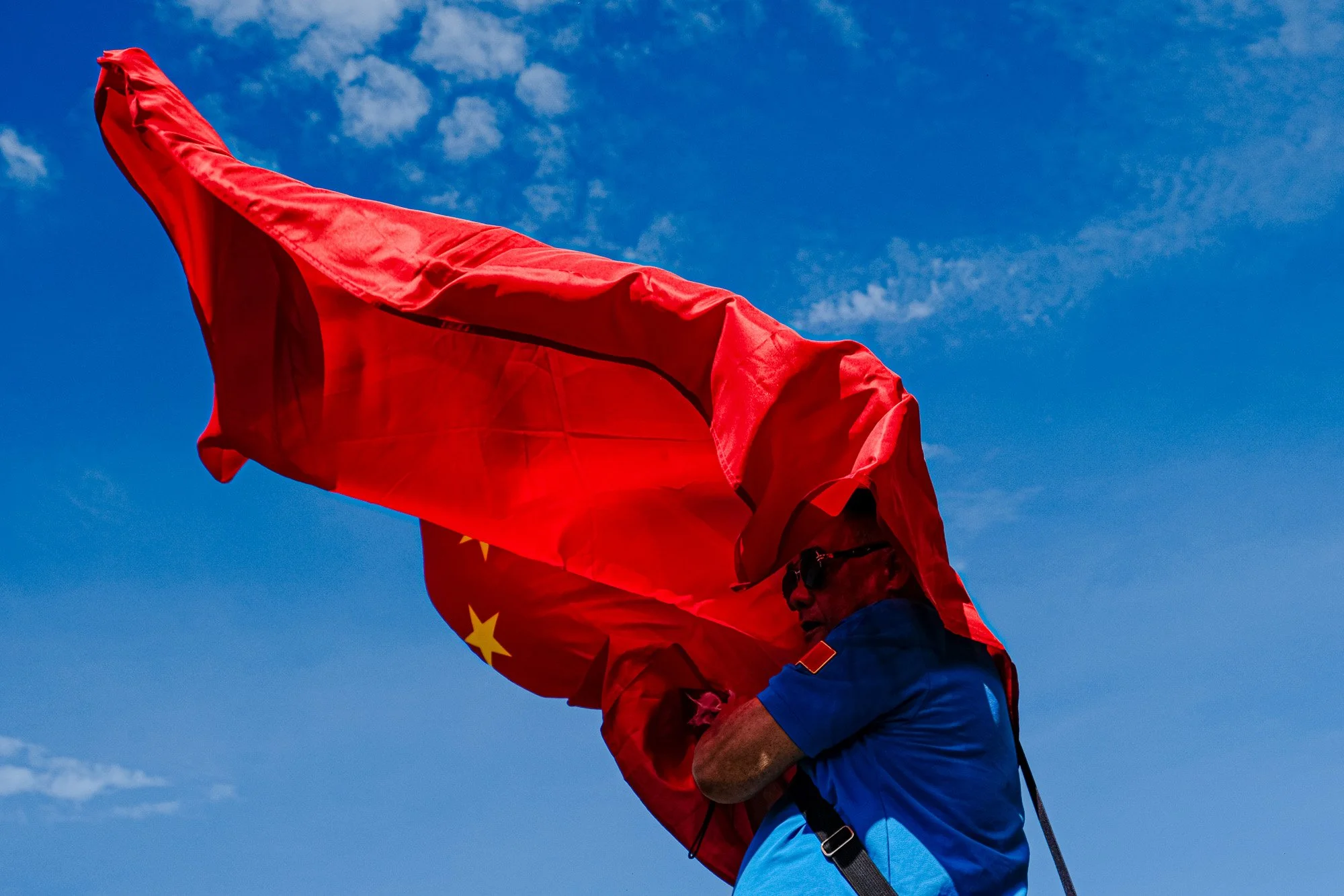 A person holding a red flag with yellow stars against a blue sky with some clouds.
