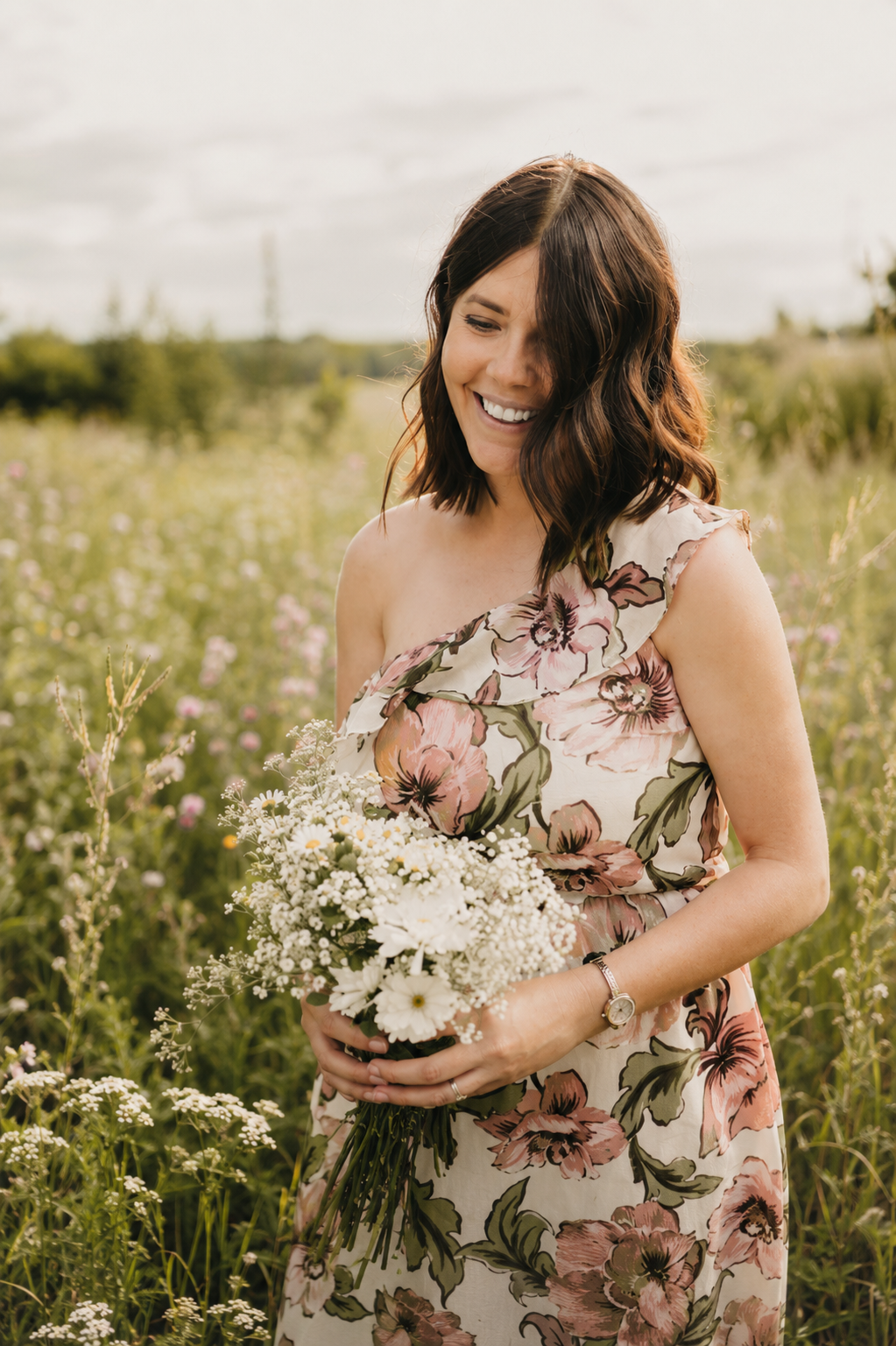 A woman in a floral dress holding a bouquet of white flowers in a field with wildflowers, smiling and looking down.