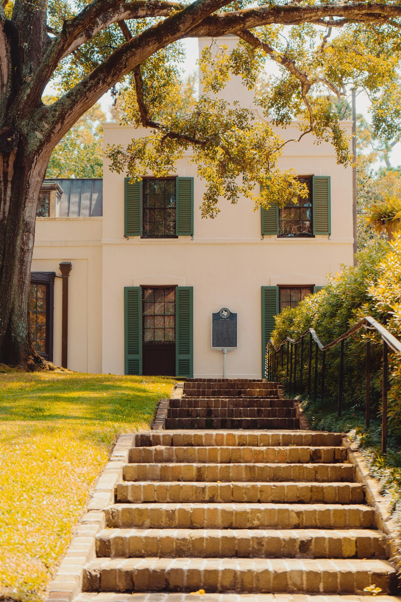 Brick stairs leading up to a white building with green shutters, surrounded by grass, trees, and shrubs on a sunny day.