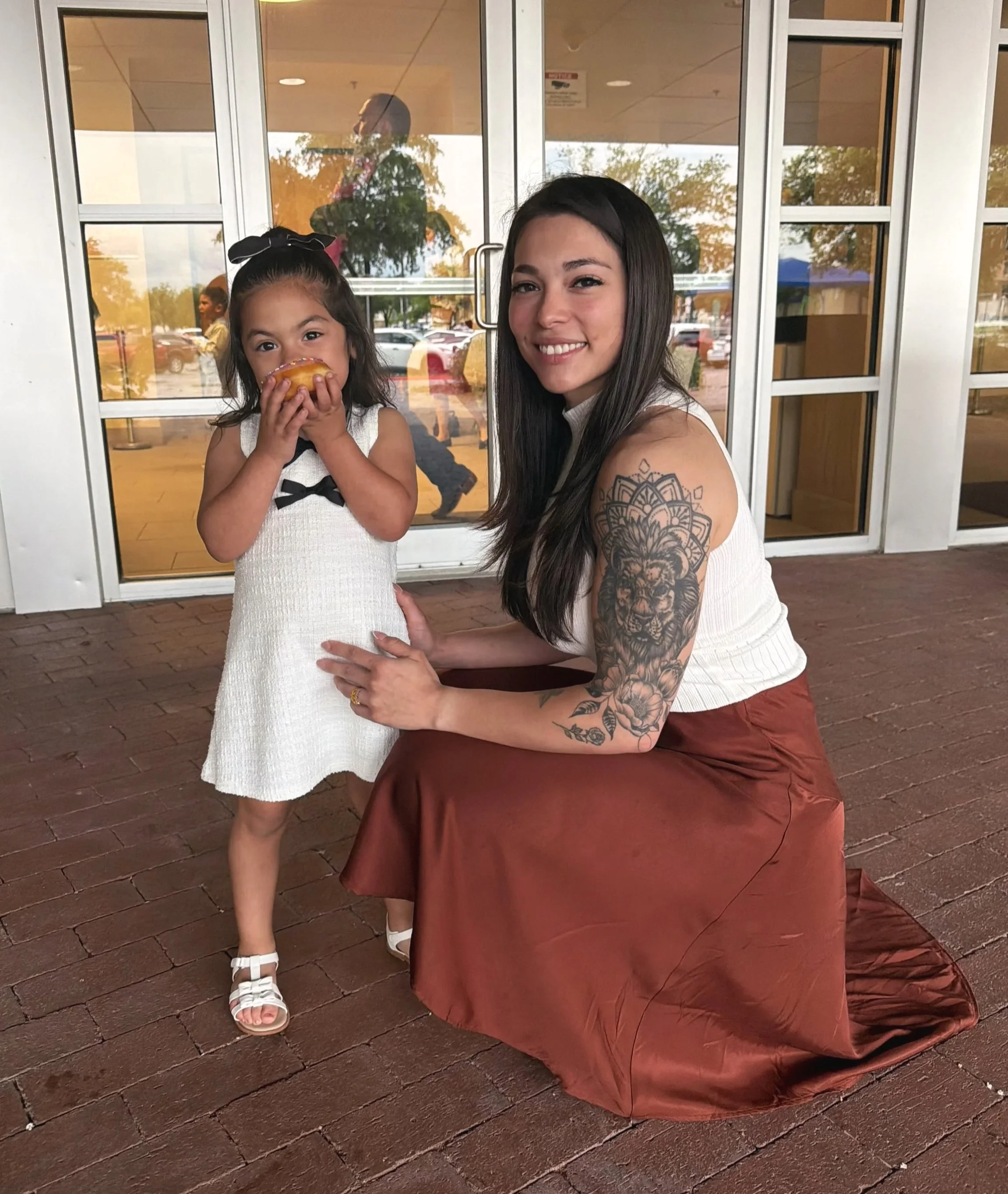 A woman with long dark hair and a tattoo on her arm kneeling on a brick floor next to a young girl in a white dress, who is eating a donut. They are outside a building with glass doors that reflect trees and parked cars.