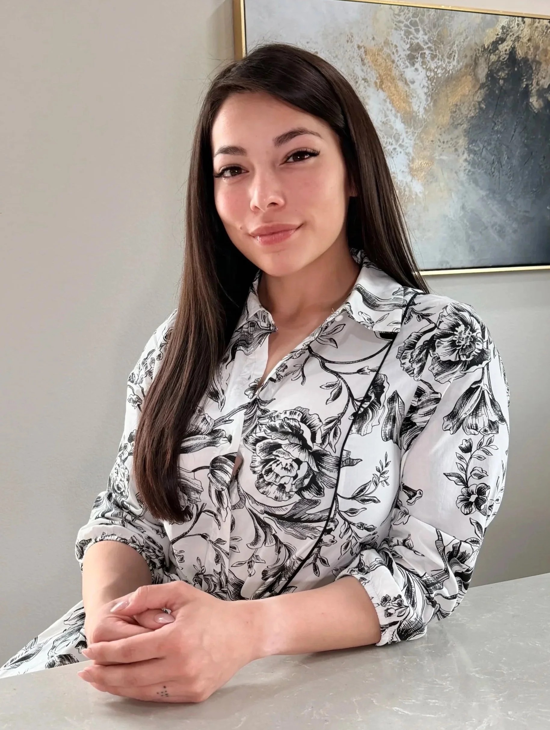 A young woman with long dark brown hair, light skin, and a slight smile, sitting at a table with her hands folded. She is wearing a white blouse with black floral patterns. In the background, there is a piece of abstract wall art with earth tones and white accents.