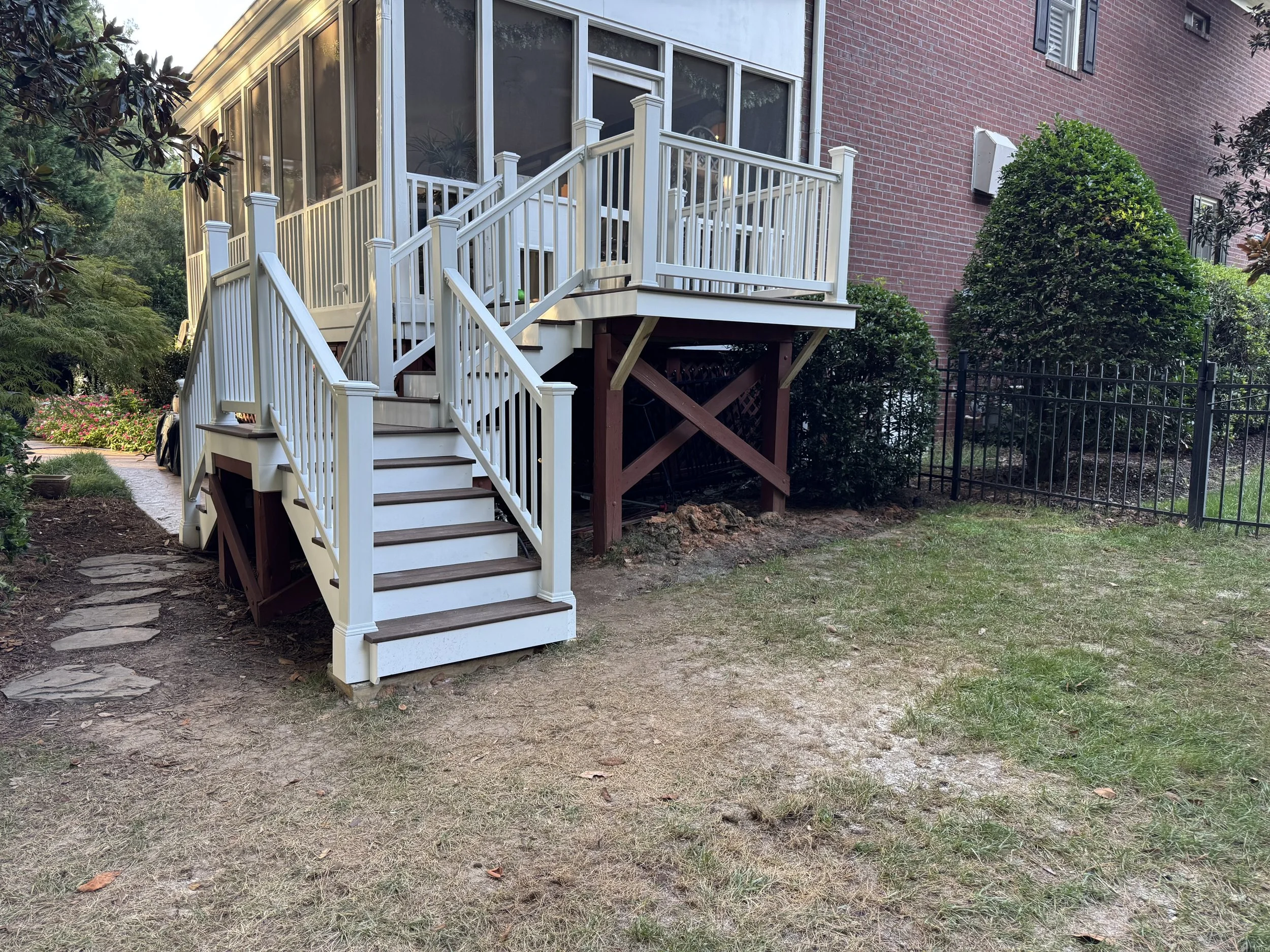Backyard with a white wooden staircase leading to a screened porch on a brick house. There are lush green bushes and trees, a gated fence, and stepping stones on the dirt path.