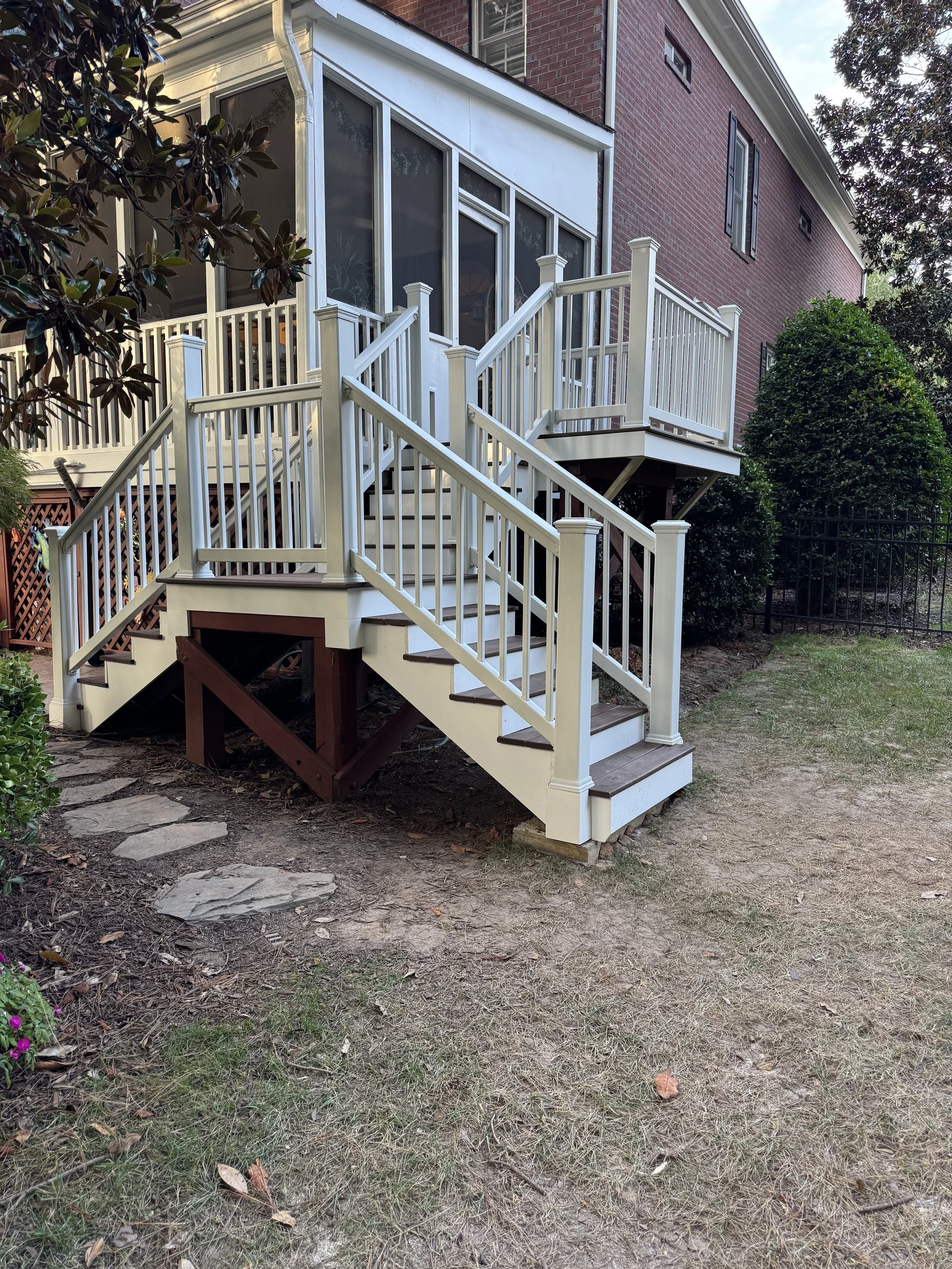 White wooden stairway with railing leading up to a screened porch on a brick house, surrounded by trees and bushes.