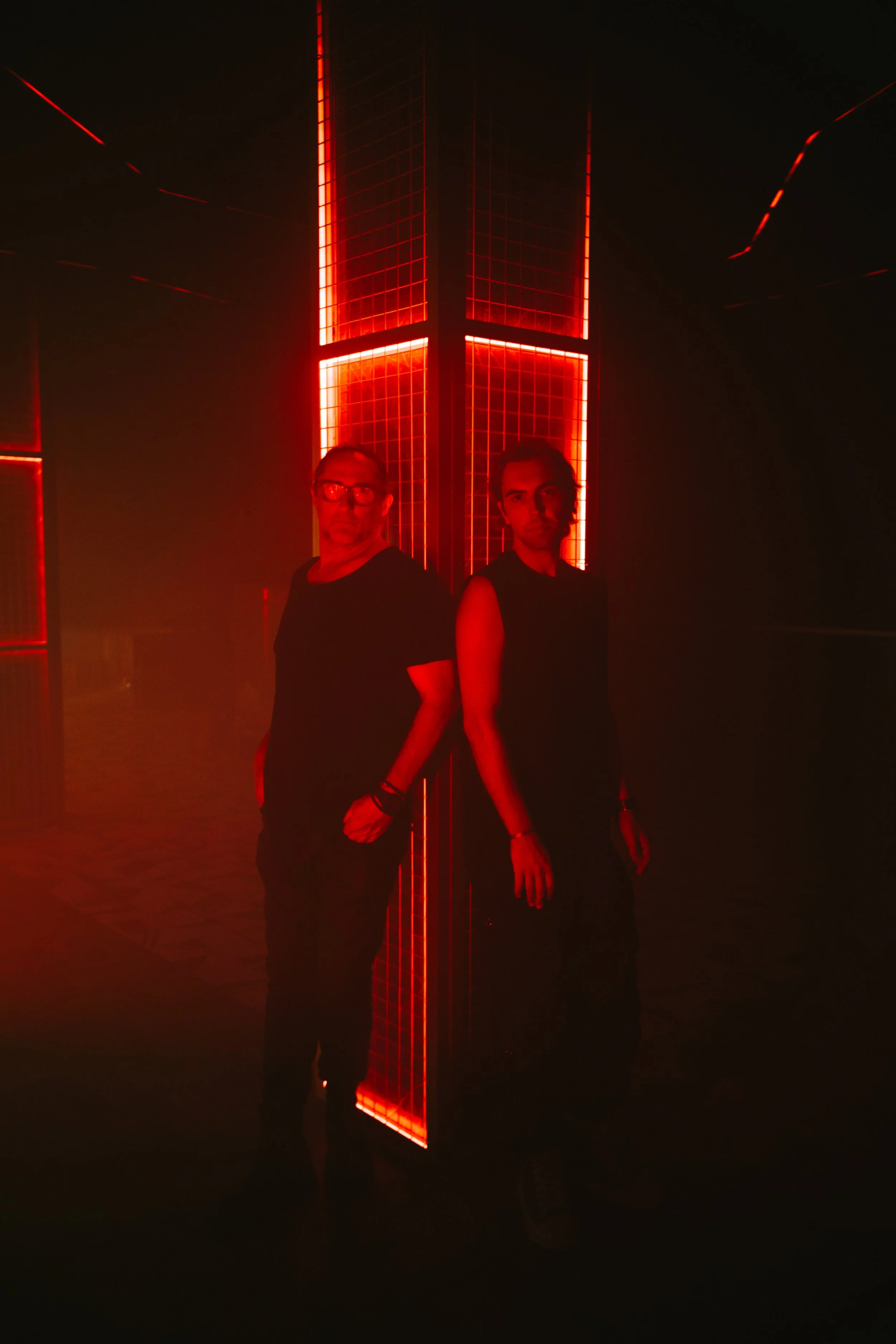 Dos personas de pie junto a una pared con luces rojas en un ambiente oscuro.