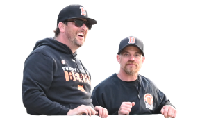 Two men wearing black hoodies and black caps with a logo, standing outdoors, smiling and looking to their left.