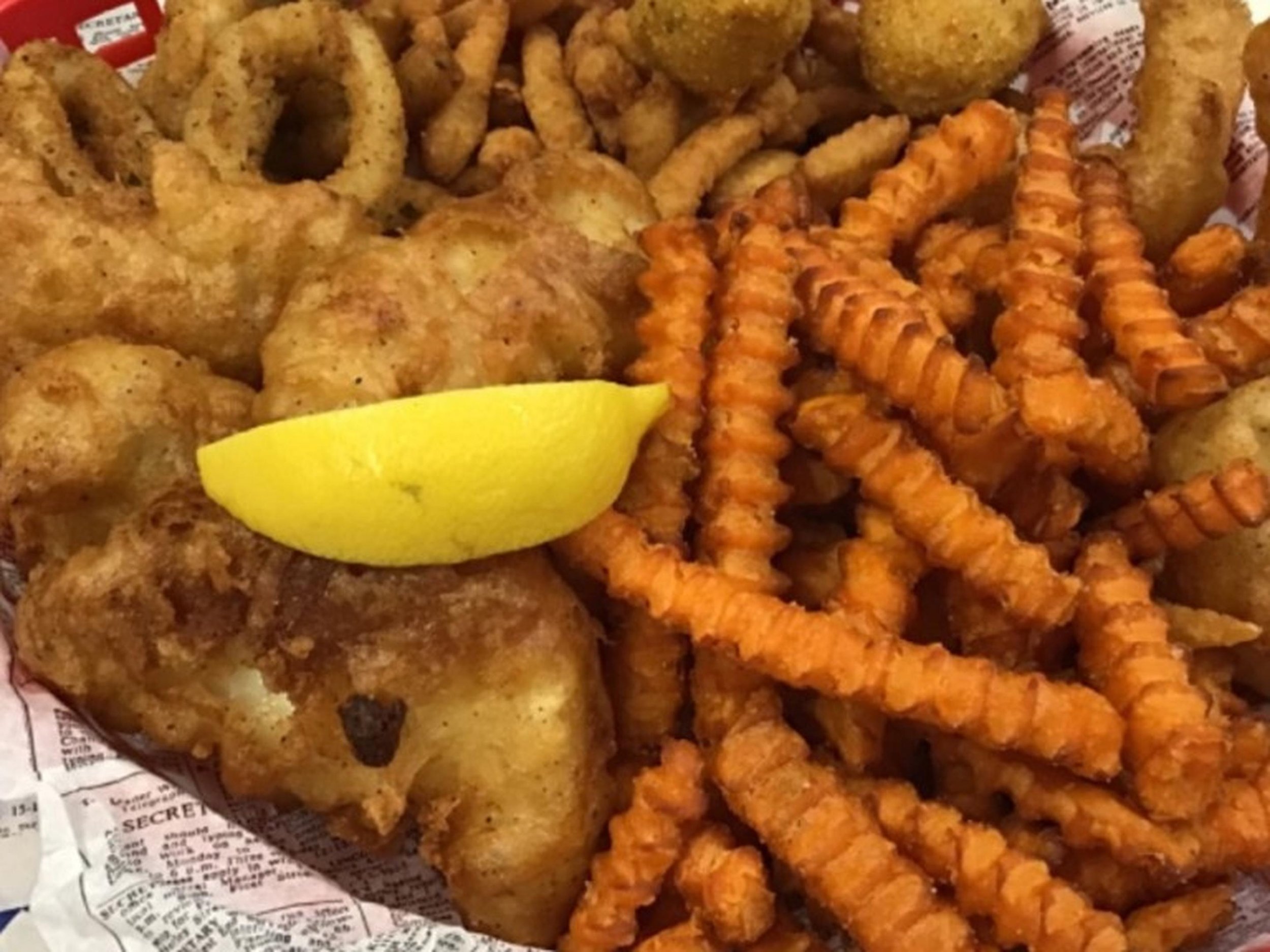 A close-up of a tray with fried chicken, curly fries, onion rings, and a lemon wedge on a newspaper-lined surface.
