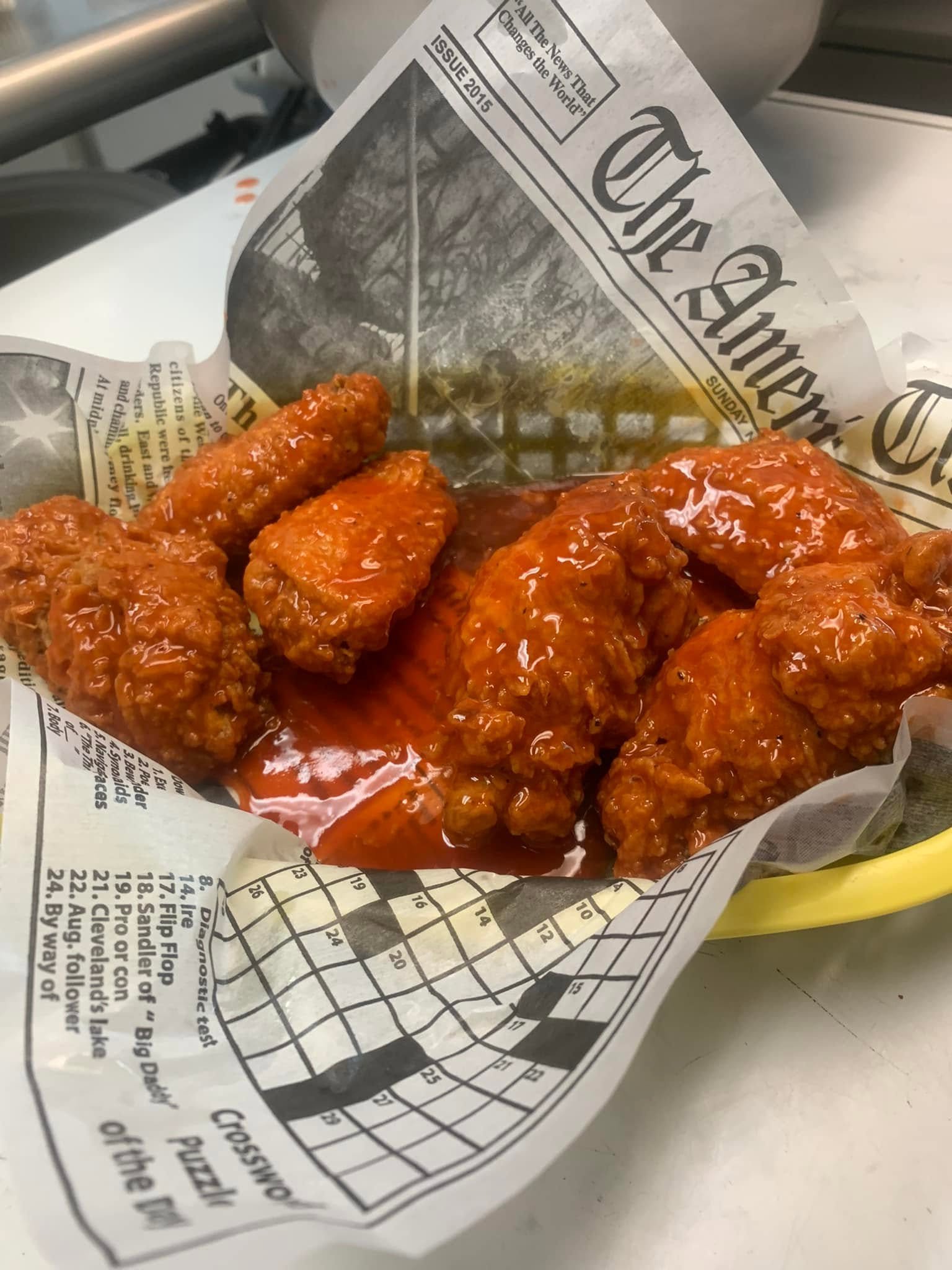 Close-up of fried chicken pieces in a red sauce, placed on paper with newspaper print and a crossword puzzle, on a metallic surface.