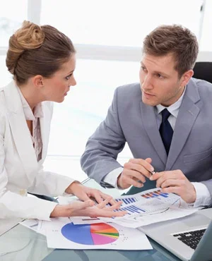 A woman and a man in business attire discussing documents and charts at a desk in an office.