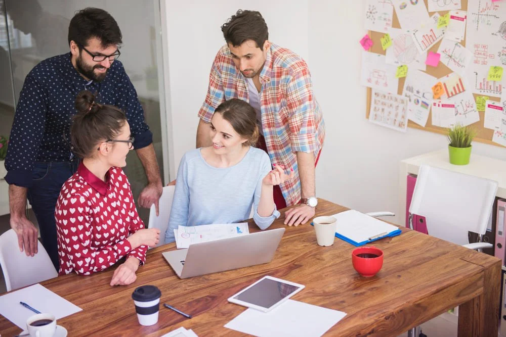 Group of five coworkers having a discussion in a modern office conference room, with laptops, papers, coffee, and a bulletin board with charts on the wall.