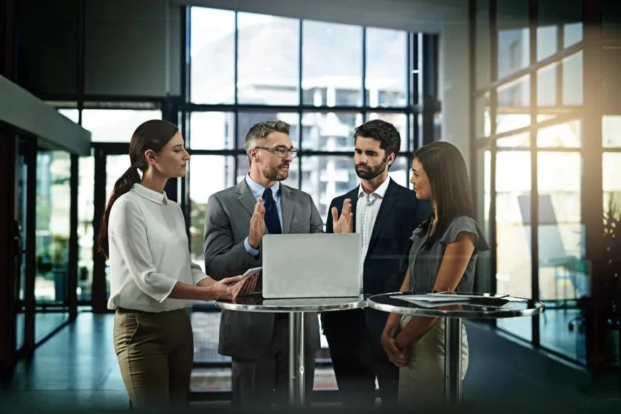 Four business professionals in discussion around a laptop in a modern office lobby with large glass windows and sunlight.