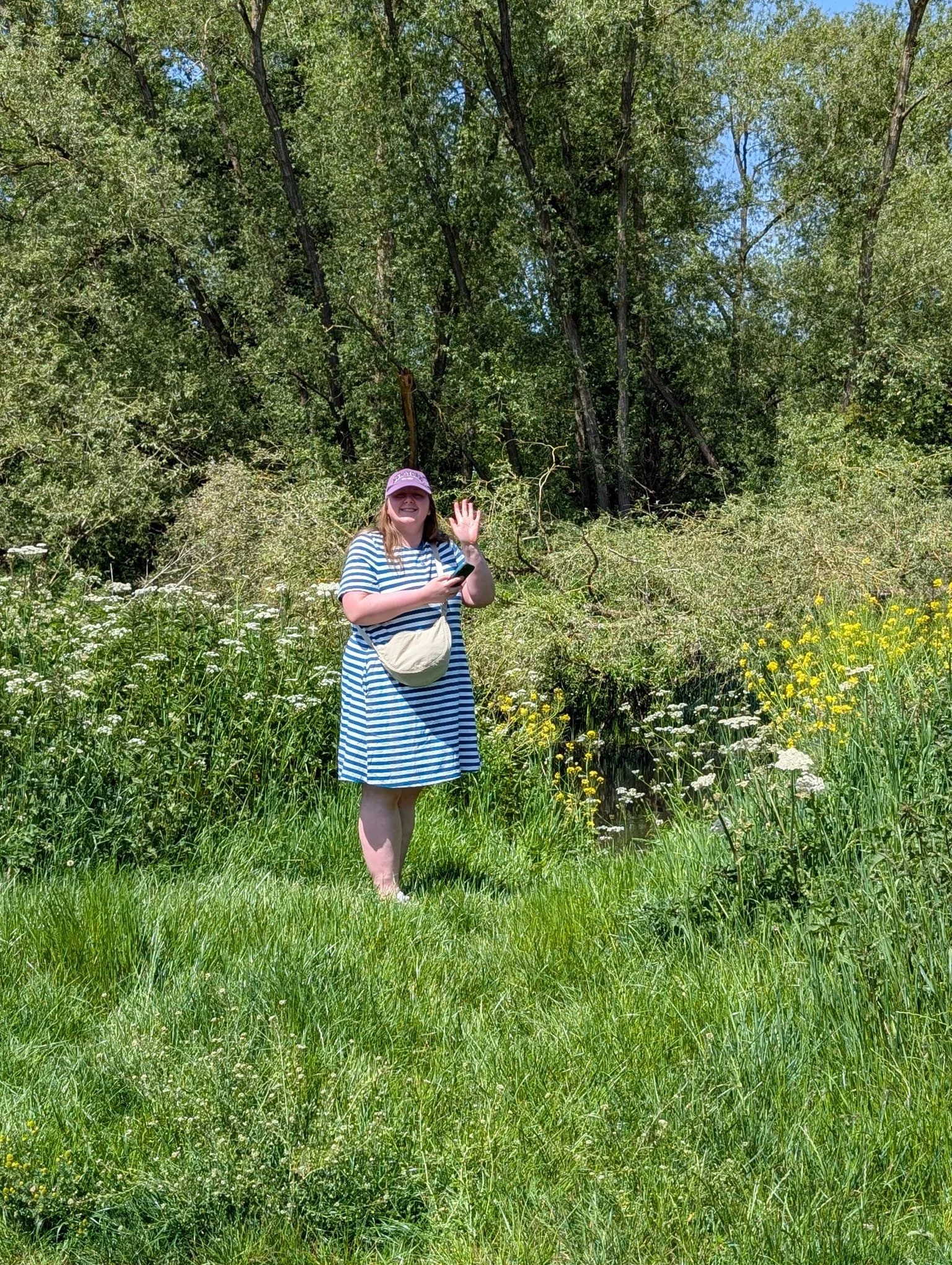 A woman wearing a striped dress, pink hat, and carrying a beige crossbody bag, standing in a lush green grassy area surrounded by wildflowers, waving and smiling at the camera.