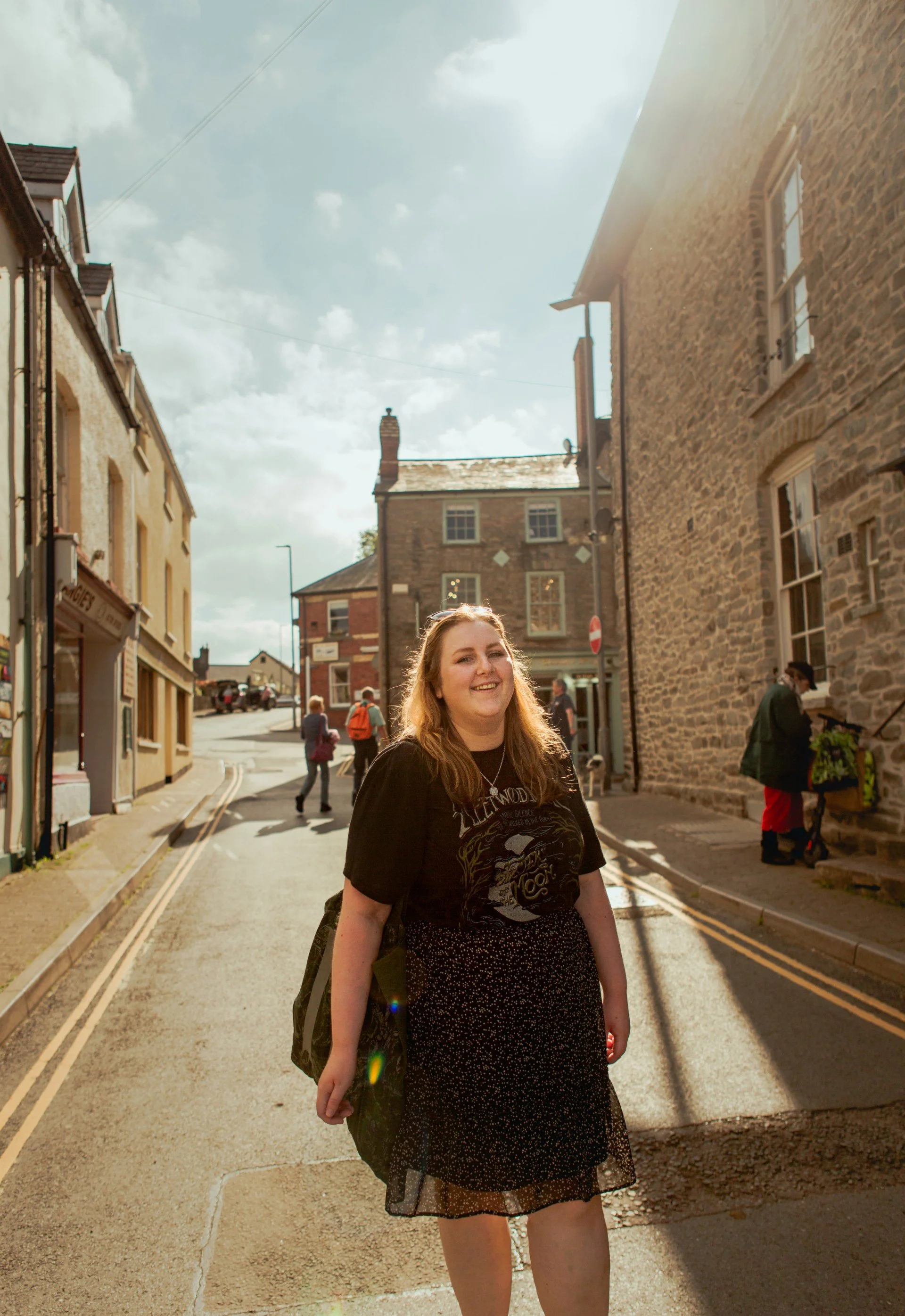A young woman with long red hair smiling, walking down a street with brick and stone buildings, in bright daylight with a partially cloudy sky.