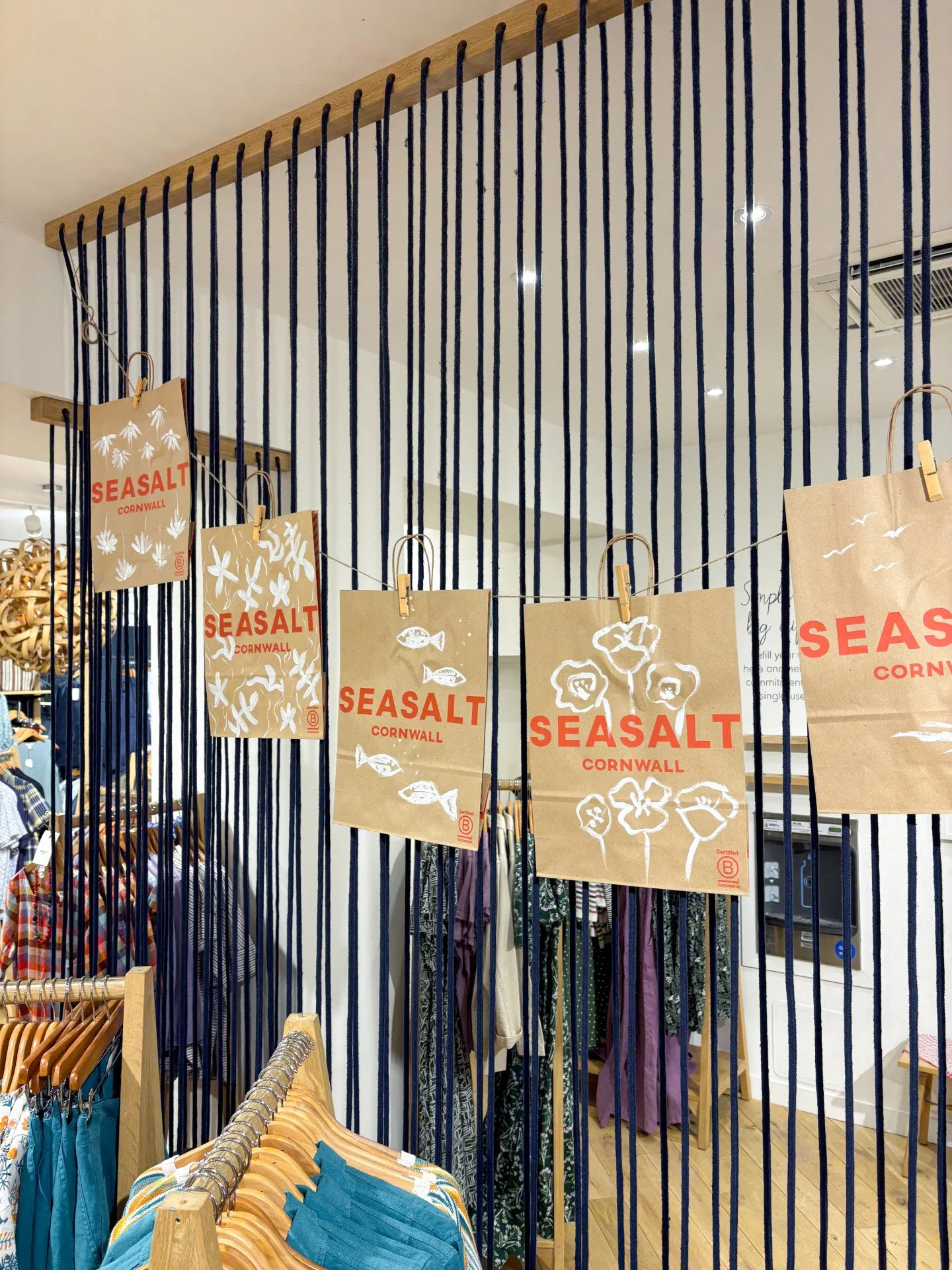 Display of illustrated brown paper shopping bags with the words 'Seasalt Cornwall' and illustrations of seashells, fish, and flowers, hanging on a string with clothespins inside a clothing store.