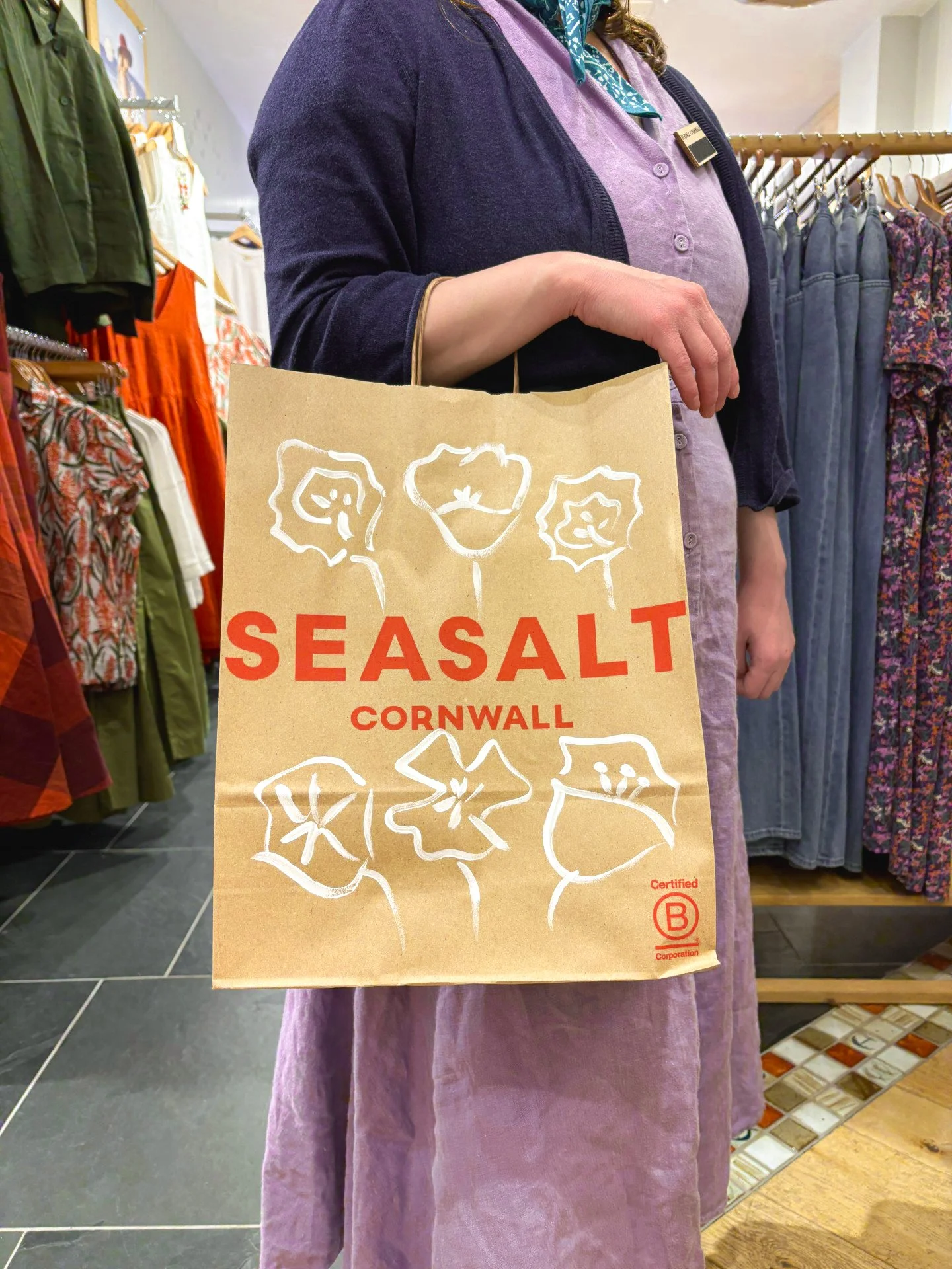 A person holding an illustrated paper shopping bag with the text 'SEASALT CORNWALL' and floral illustrations, standing inside a clothing store.