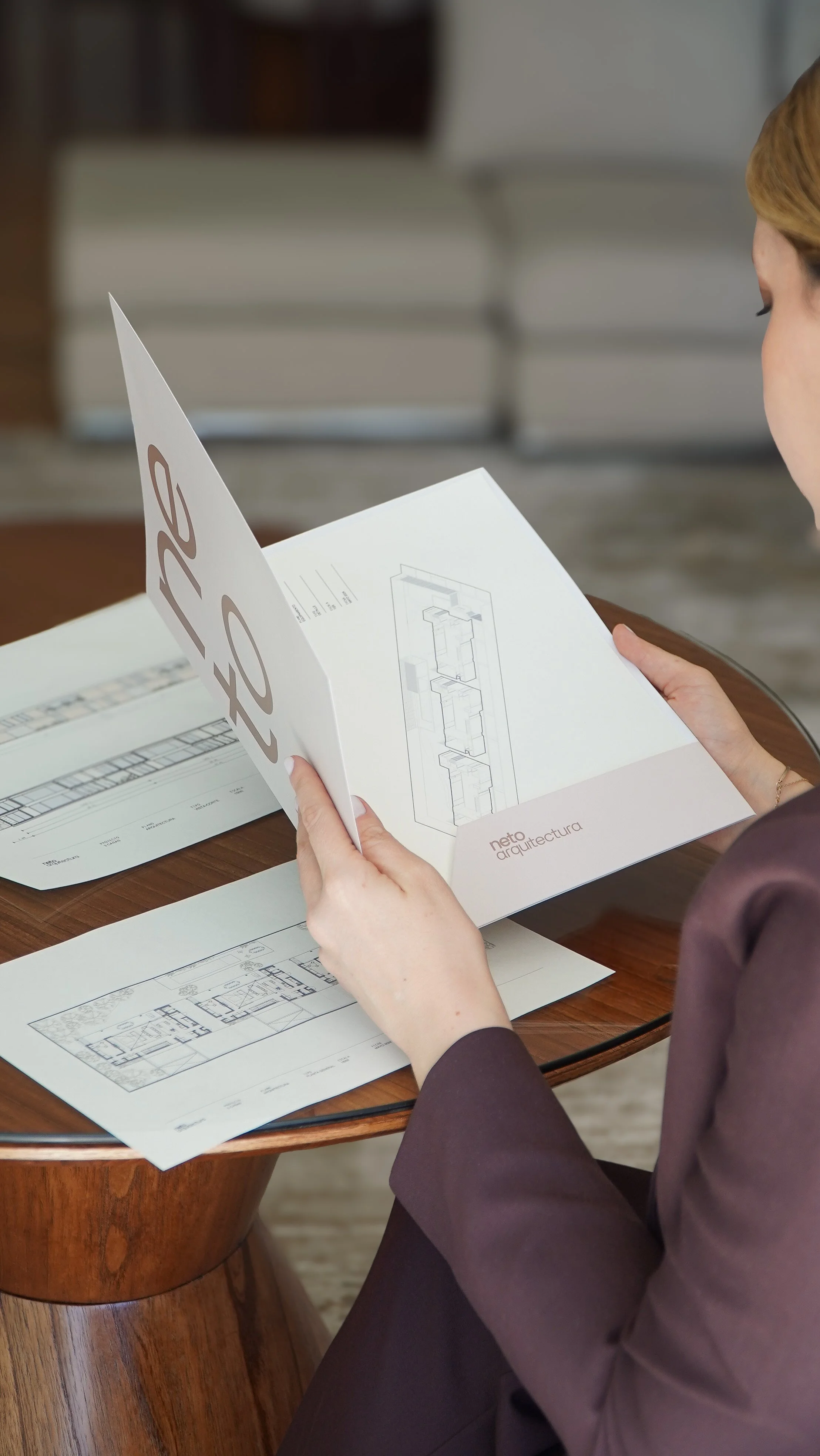 Persona revisando un folleto de arquitectura en una mesa de madera, con planos y dibujos técnicos.