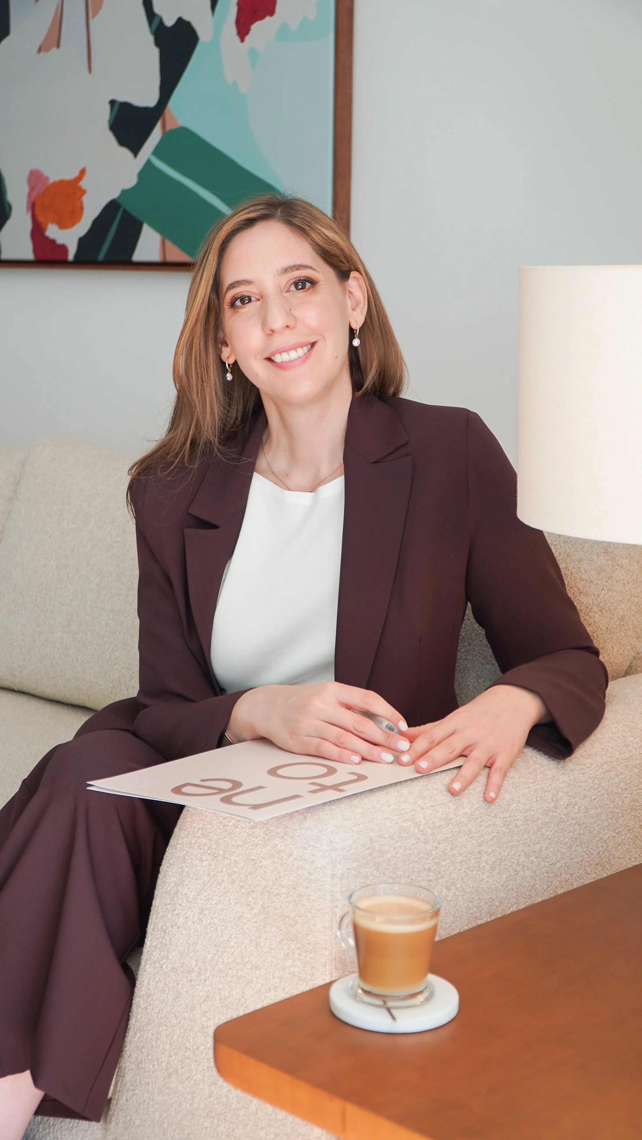 Una mujer sonriente con vestimenta formal sentada en un sillón, frente a una mesa con una taza de café, en un entorno de oficina o sala de reuniones.