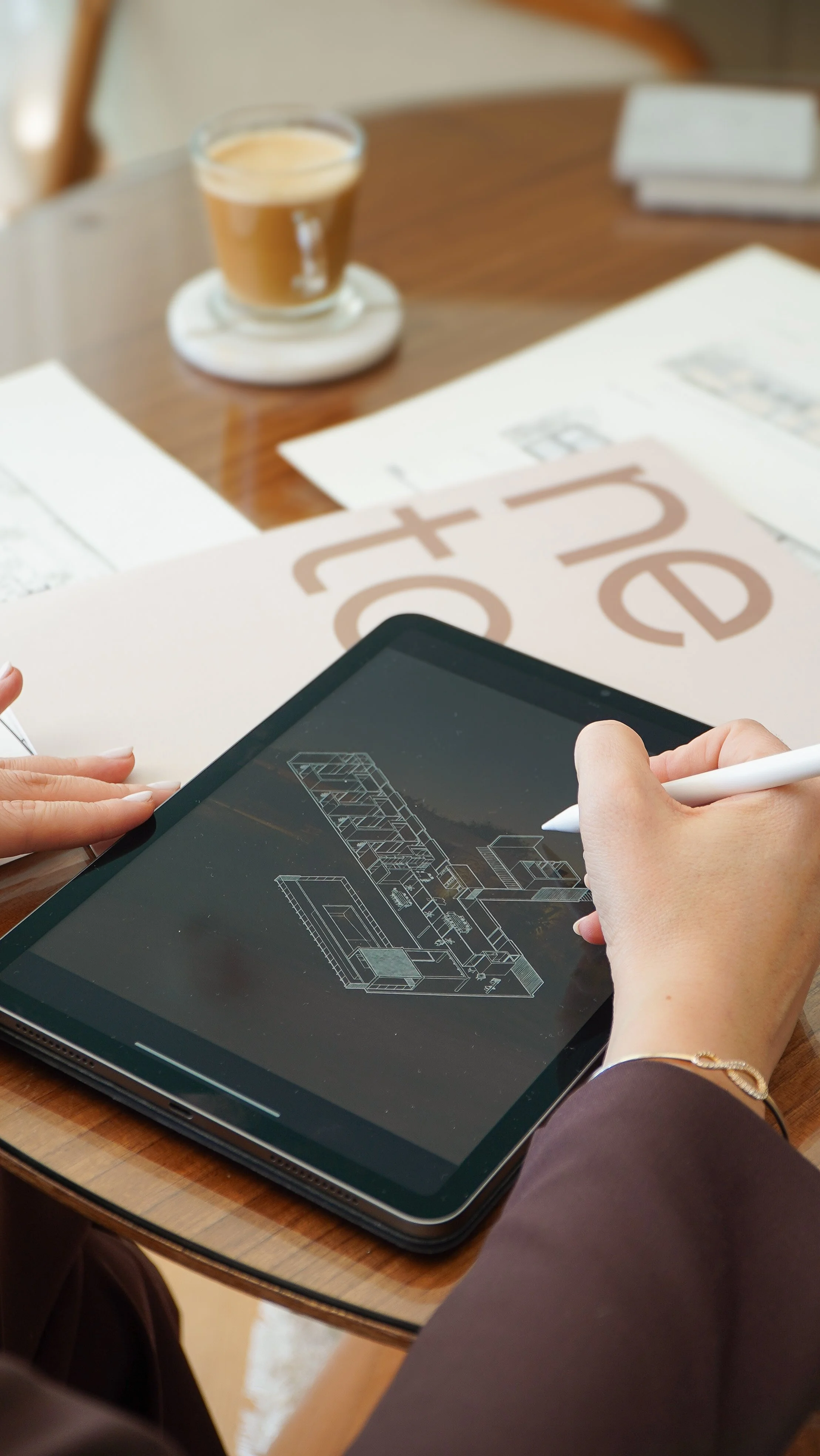 Persona usando una tableta con un diseño arquitectónico en pantalla, en una mesa de madera con papeles y un café