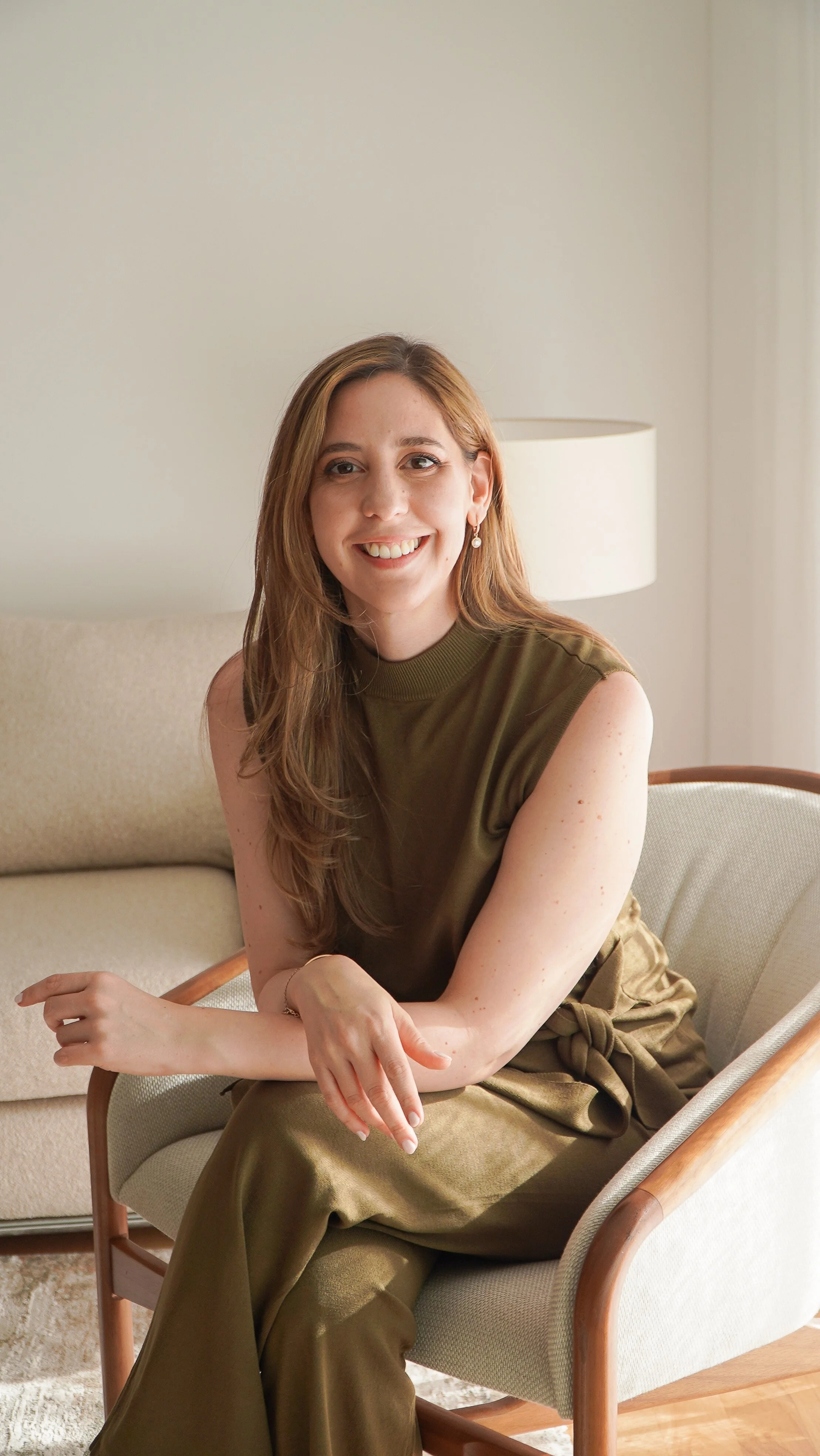 Mujer con cabello castaño claro y sonrisa, vestida con blusa sin mangas verde oliva y pantalones a juego, sentada en una silla con marco de madera en una habitación con pared blanca y lámpara de mesa cremosa.
