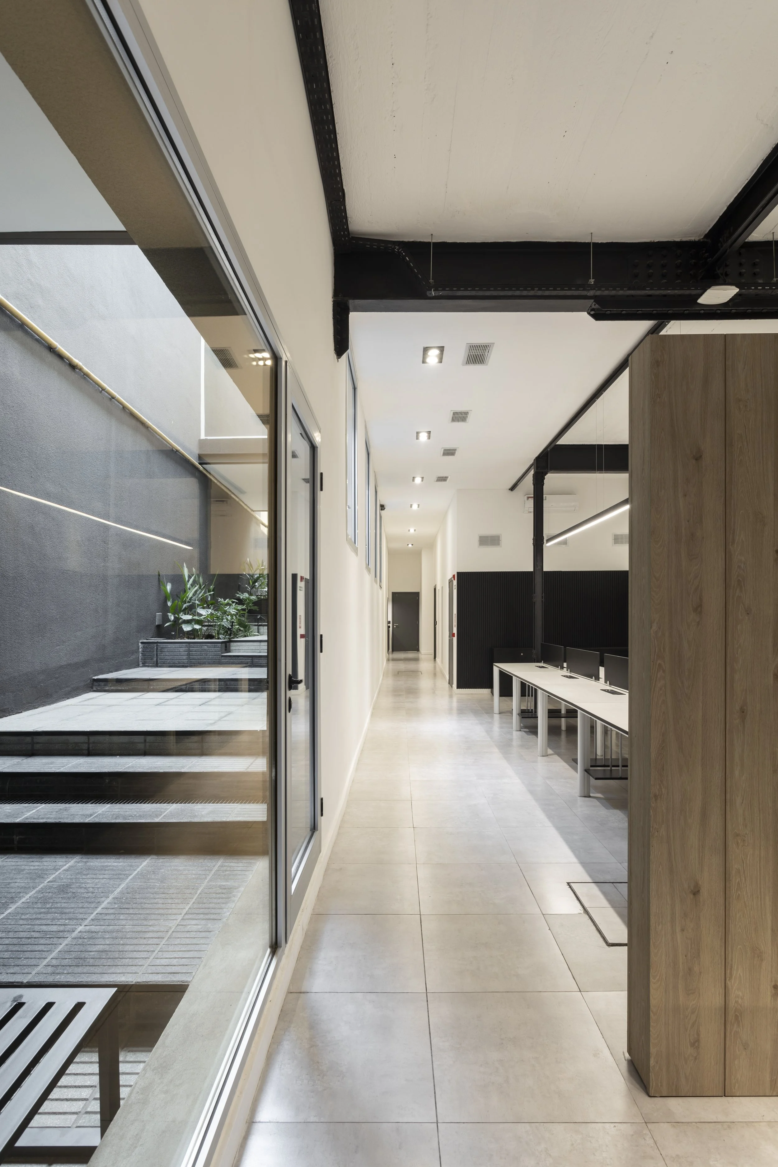 Pasillo de oficina con ventanas de vidrio, mesas de trabajo con computadoras, pared de madera y luces empotradas en el techo.