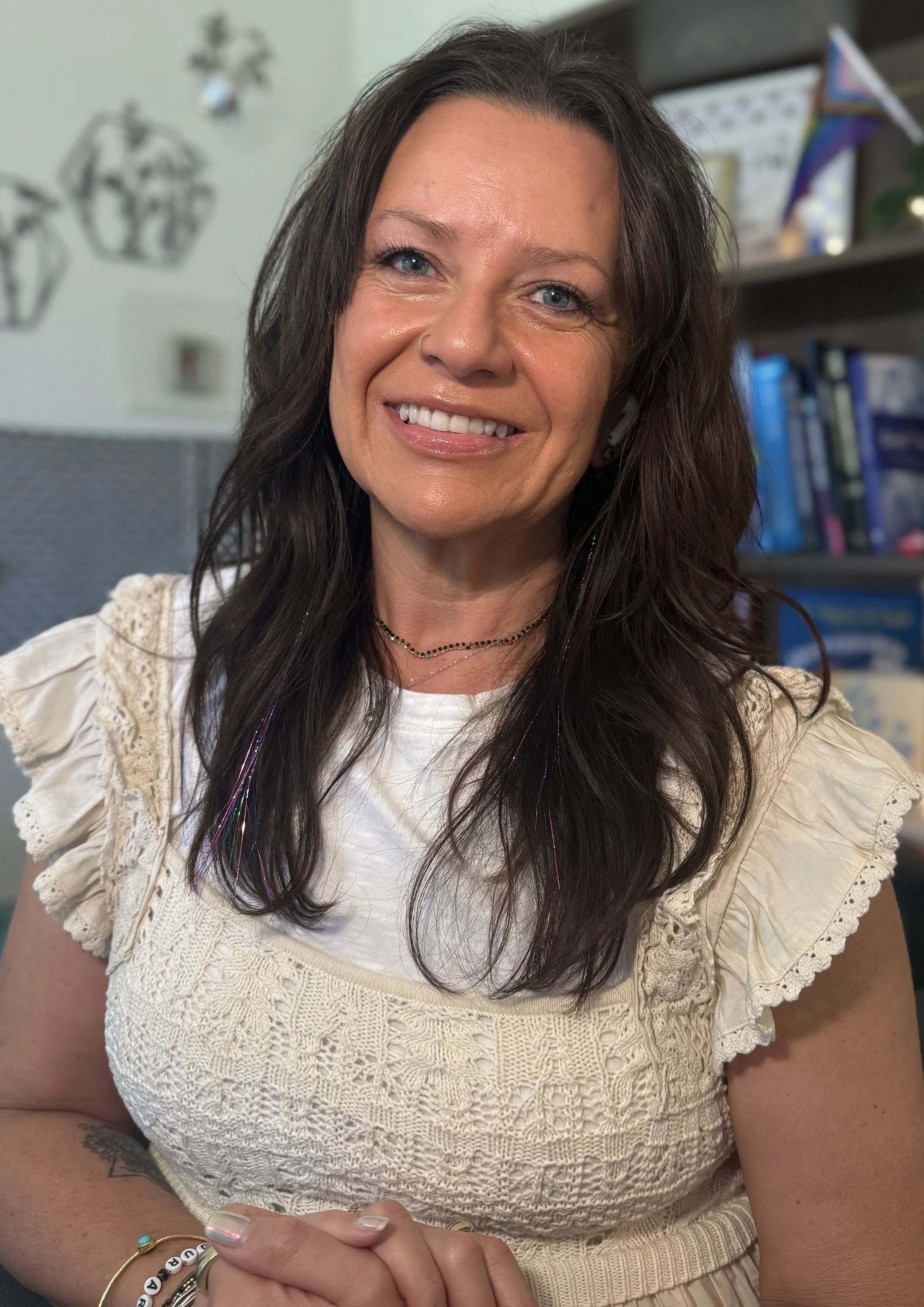A woman with long dark hair, blue eyes, and a nose piercing smiling at the camera. She wears a cream-colored lace top, layered necklaces, and bracelets, with a colorful beaded bracelet on her wrist. Background includes bookshelves and decorations.