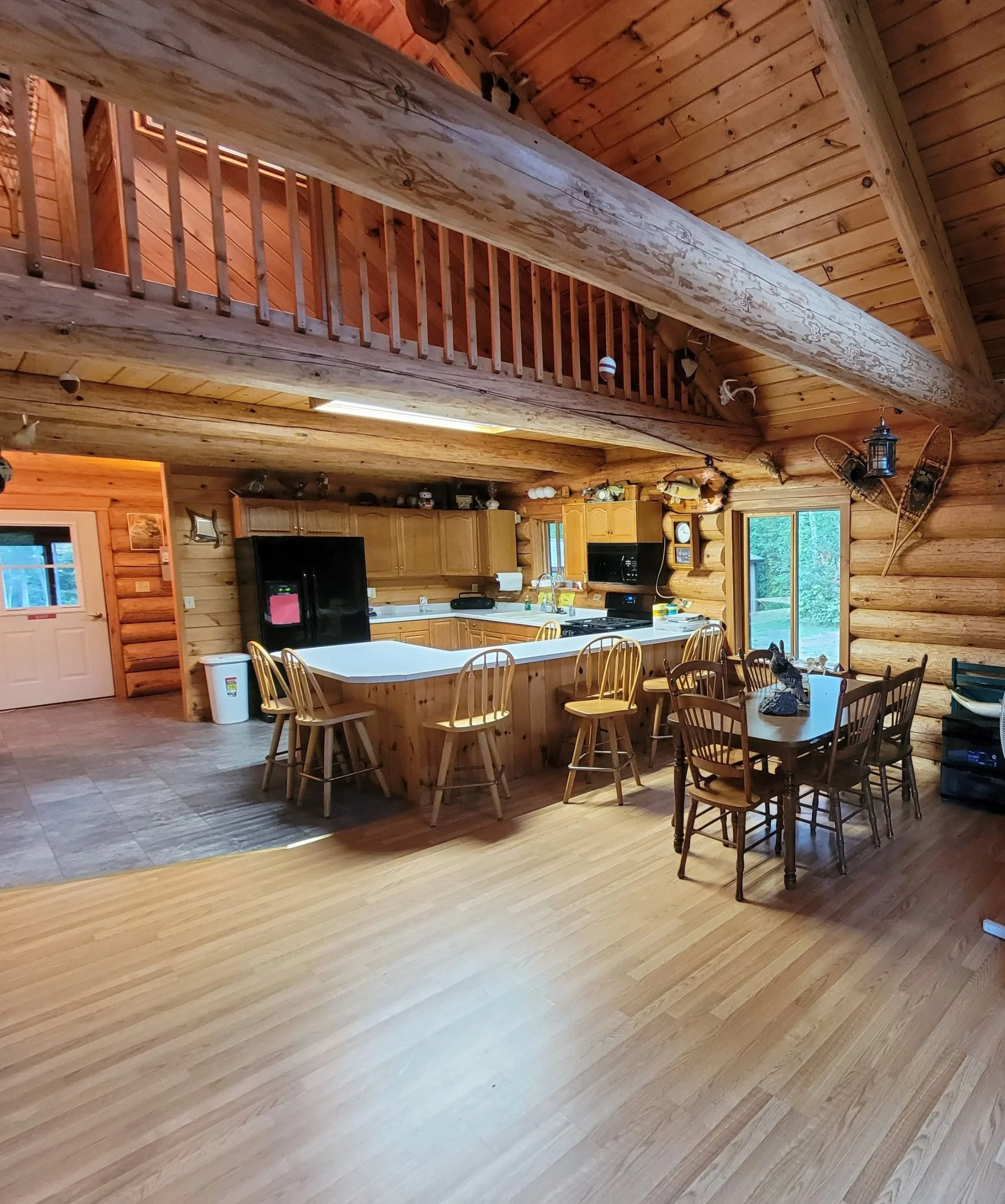 Interior view of a log cabin kitchen and dining area with wooden walls and ceiling, a large kitchen island with wooden chairs, a black refrigerator, and a dining table with matching wooden chairs. Large windows and a sliding glass door provide natural light and outdoor views.