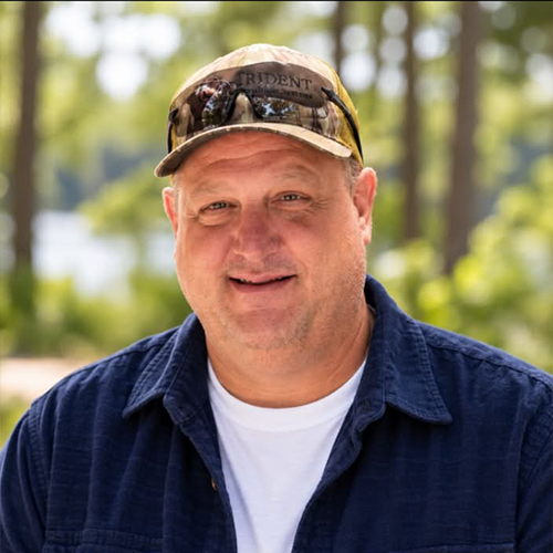 Man wearing a black and orange Carolina hat, sunglasses, camouflage jacket, standing outdoors in a wooded area during daytime.