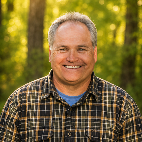 Smiling middle-aged man with gray hair, wearing a plaid shirt, outdoors with trees in background.