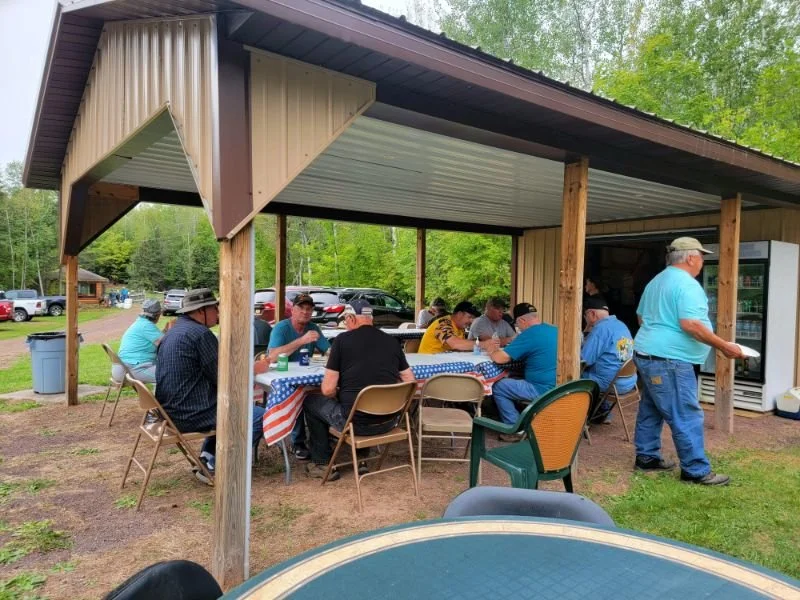 People gathered under a pavilion having a meal with a decorated tablecloth, outdoor setting with trees and parked cars in the background.