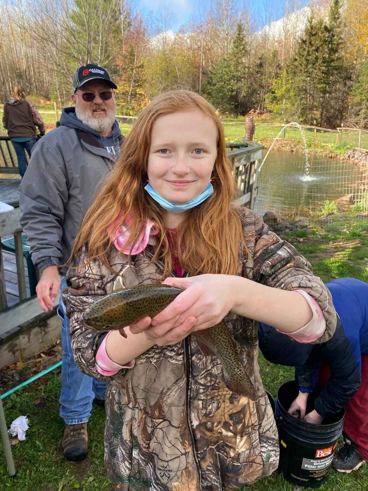 A young woman with red hair and a camouflage jacket holding a fish, smiling at the camera, outdoors near a pond with a fountain, surrounded by trees with autumn foliage, and people in the background.