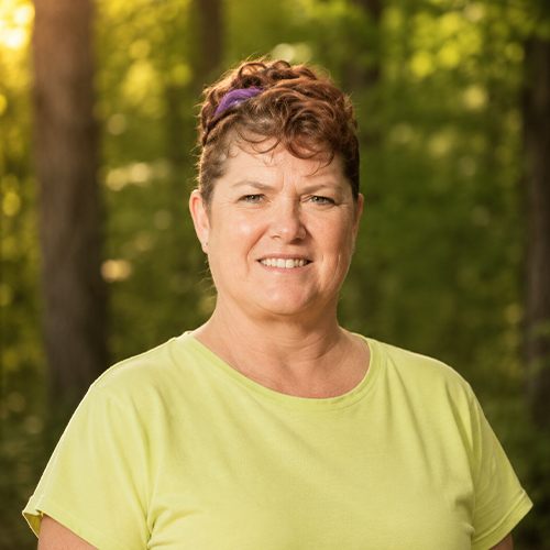 A woman with short curly brown hair, wearing a yellow t-shirt and a purple headband, smiling outdoors with green trees in the background.