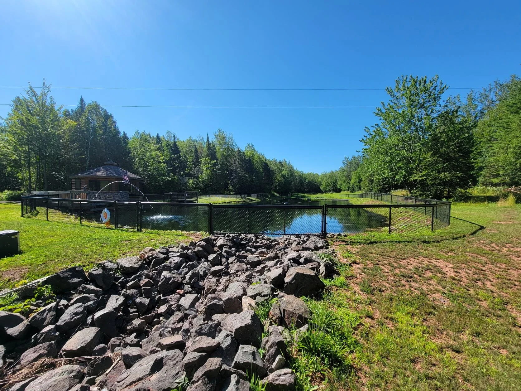 A fenced pond with a small gazebo on the left, surrounded by trees, rocks, and grassy areas on a bright sunny day.
