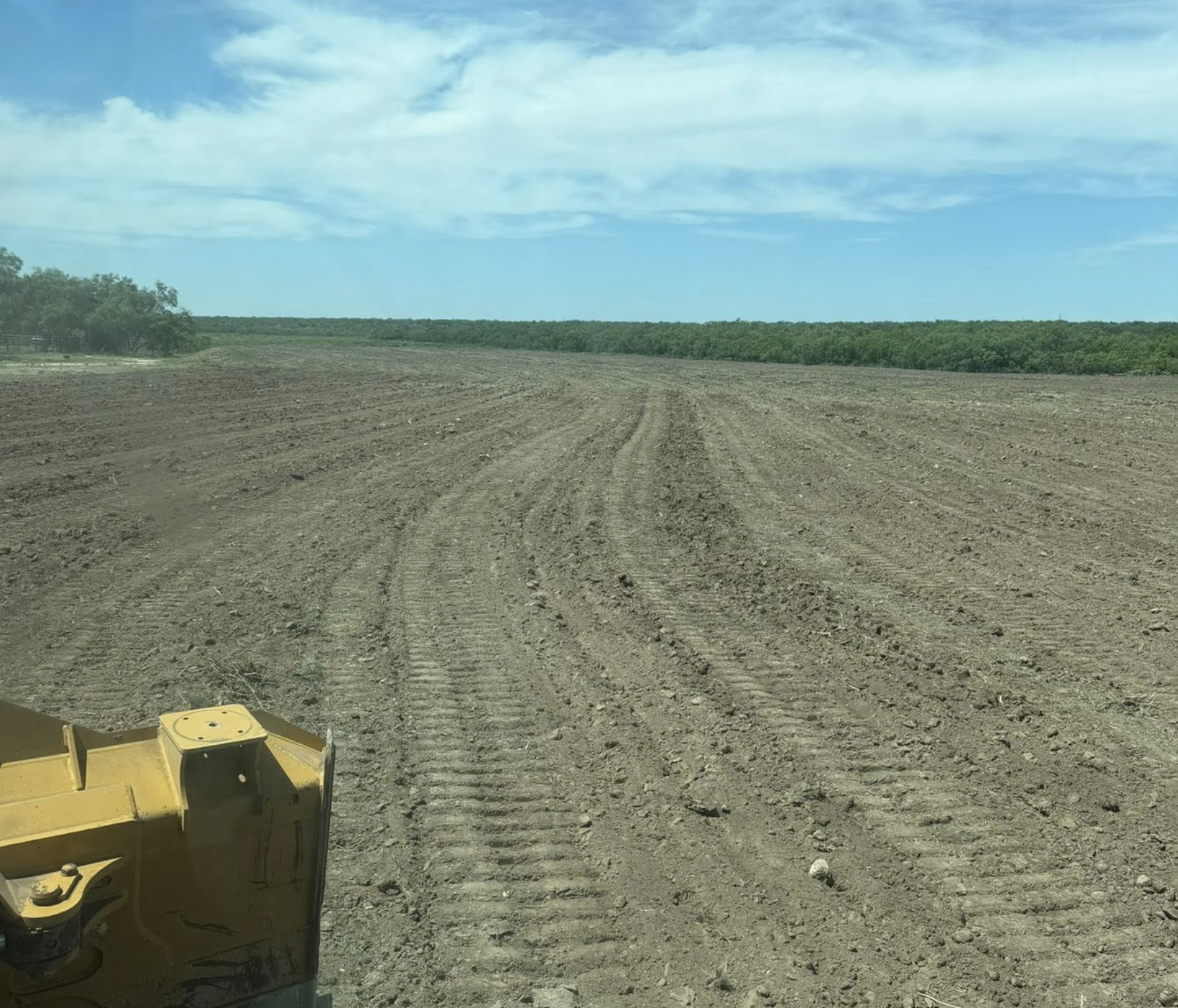 A cleared, plowed field extending to a distant tree line under a partly cloudy blue sky, with some construction machinery visible at the bottom left corner.